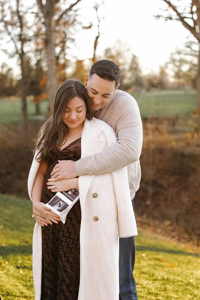 Golden Hour Fall Pregnancy Announcement Photos of an expecting couple wrapped in a cozy embrace, with an ultrasound photo in hand and warm fall sunlight creating an intimate, heartfelt moment.