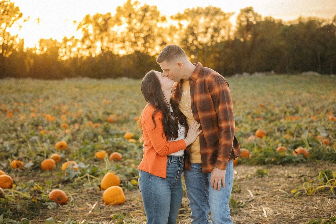 Couple sharing a kiss in the pumpkin patch at Dull’s Pumpkin Harvest, wearing orange autumn layers as golden sunlight illuminates the distant trees