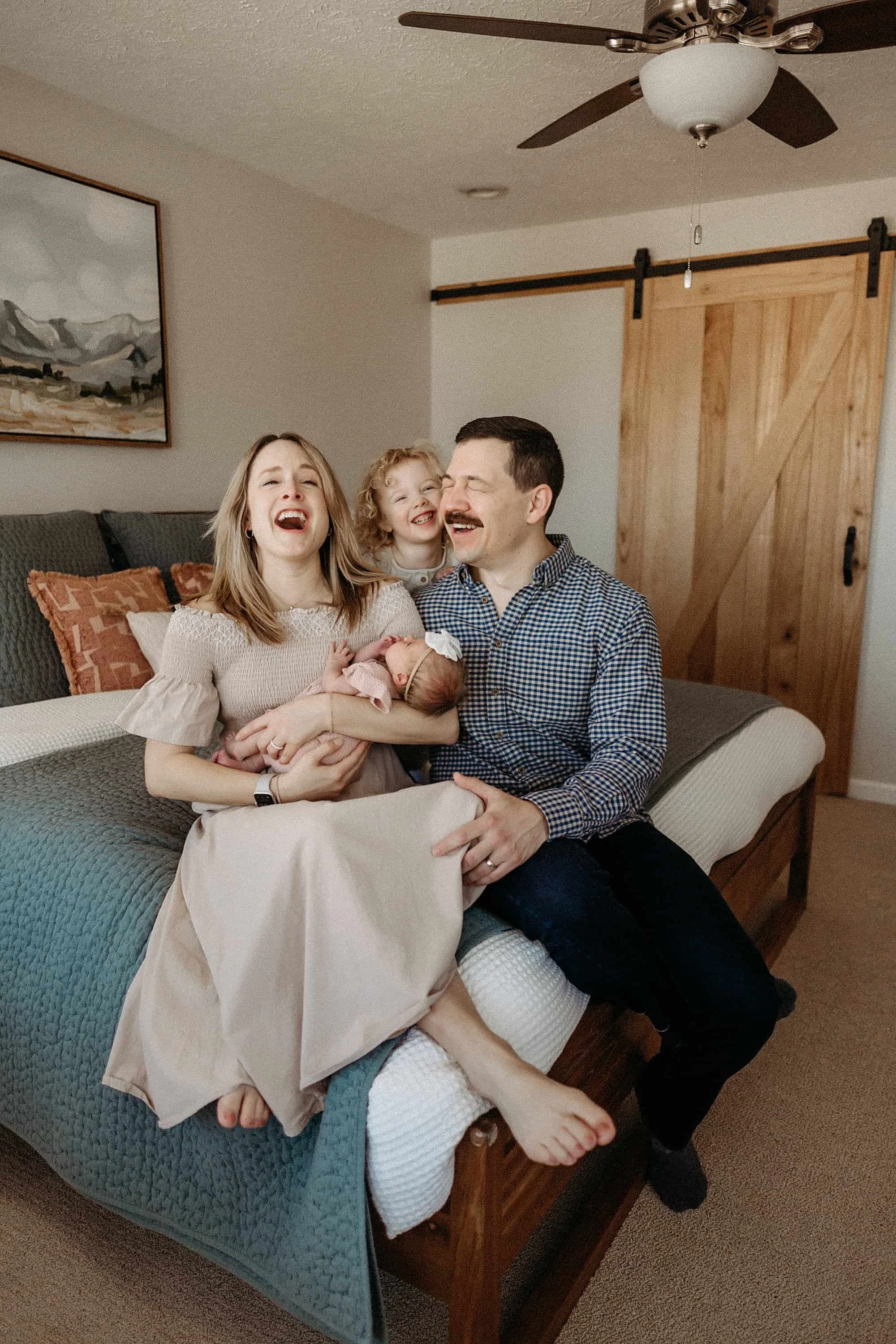 An Indianapolis family of four with a newborn sitting on a bed, laughing and smiling. The mother is holding a newborn baby, the father is sitting next to her, and a young daughter stands behind them..