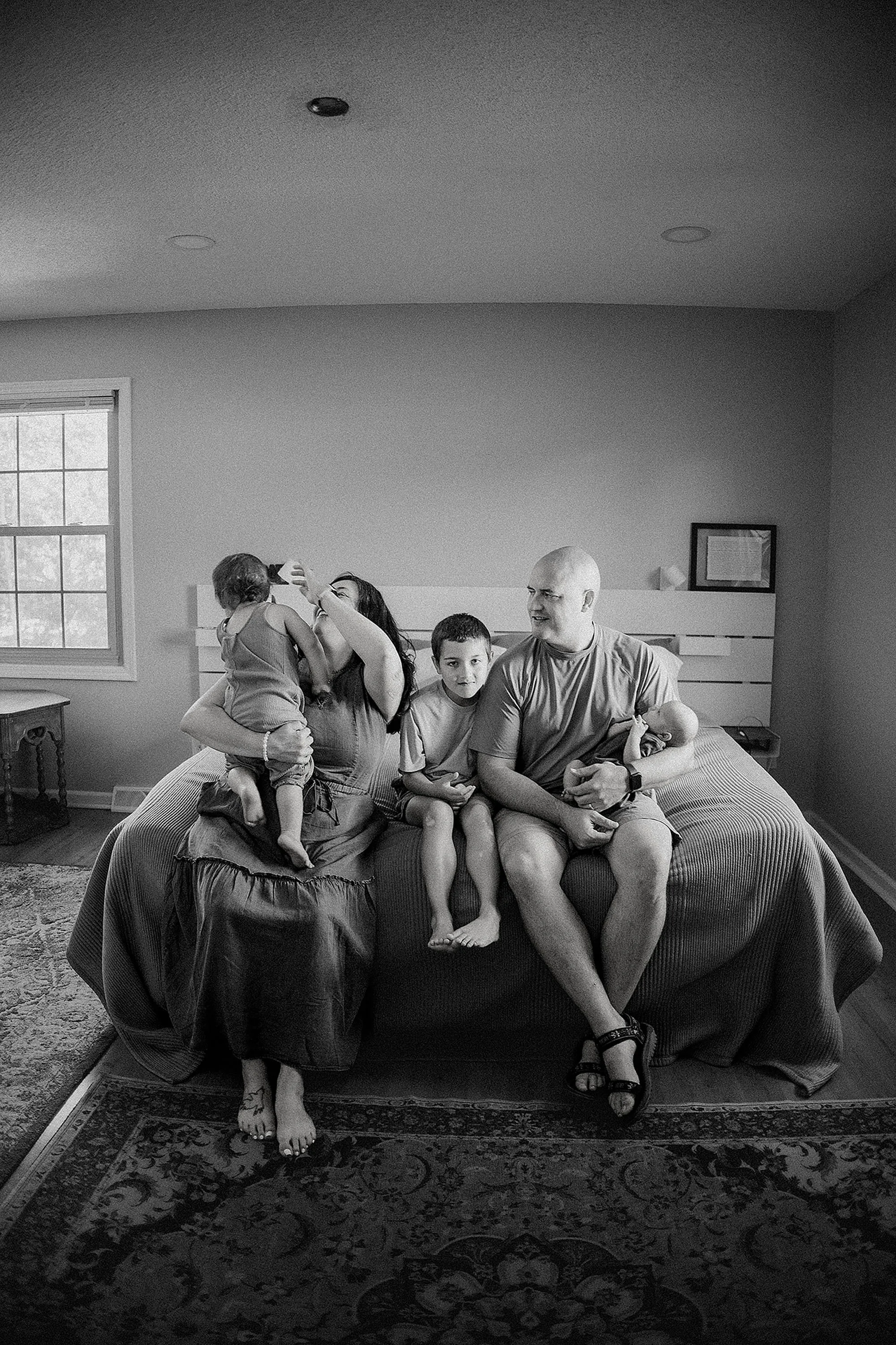 A family of five sitting on a bed in a bedroom. An adult woman is holding a young child and feeding them, two children are sitting on the bed, and an adult man is holding a baby.