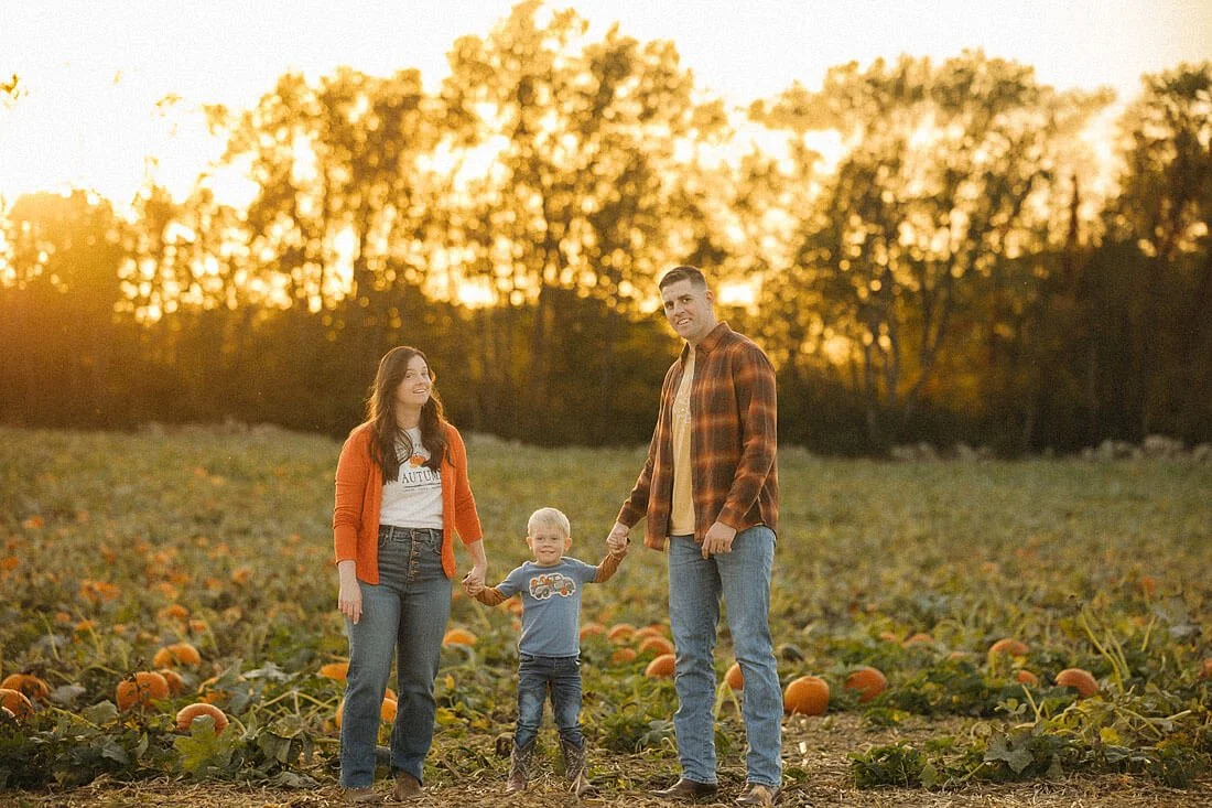 Family of three holding hands among orange pumpkins at Dull’s Pumpkin Harvest, with golden evening sunlight filtering through the trees behind them
