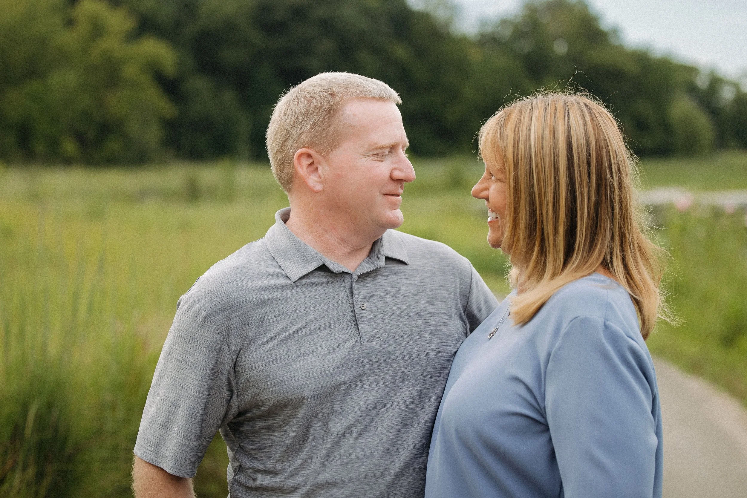 A man and woman smile at each other in a sunny field, capturing a joyful moment for their extended family photo