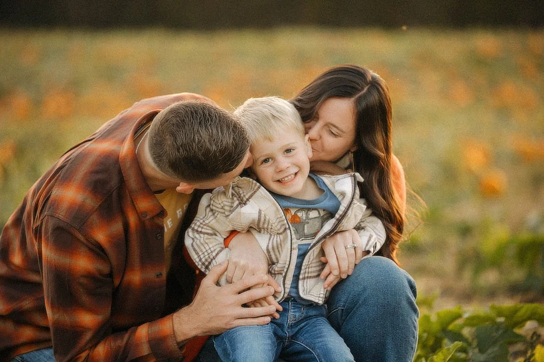 Parents kiss their smiling little boy at Dull’s Pumpkin Harvest, cuddling him close with orange pumpkins softly blurred in the background