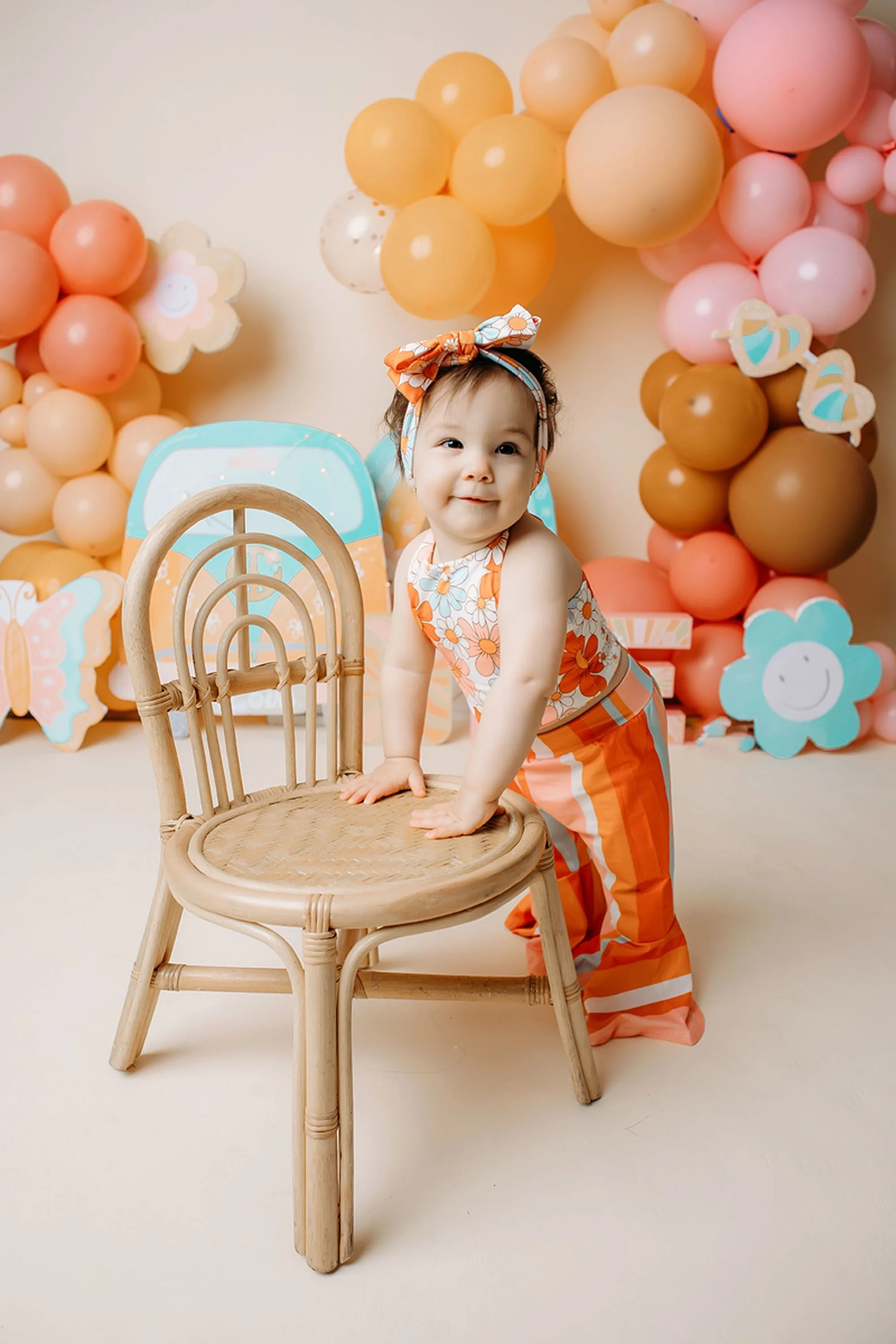 Baby girl in a retro floral halter top and orange striped pants, wearing a matching bow headband, standing and holding onto a small rattan chair with balloon garland behind her