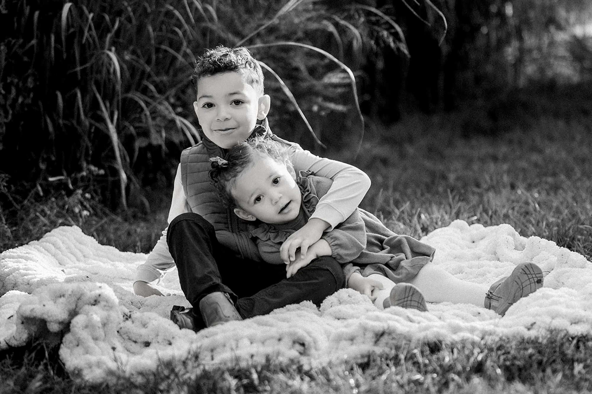 Black-and-white portrait of a young brother hugging his little sister on a chunky knit blanket in the grass — Virginia B. Fairbanks Art & Nature Park family photos capturing a timeless sibling moment