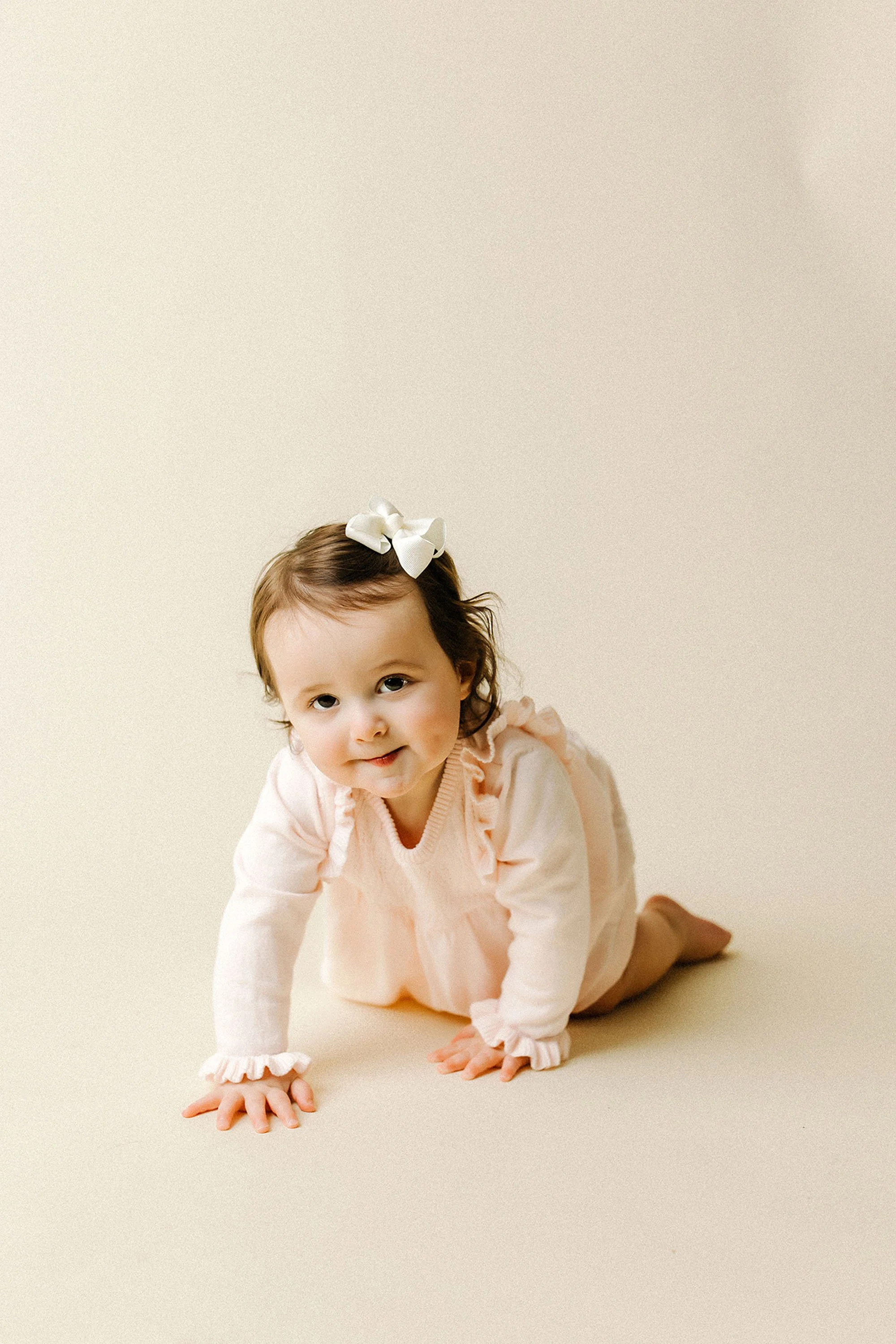 A baby girl in a pink dress crawls on the floor during her first birthday milestone session in Westfield, Indiana