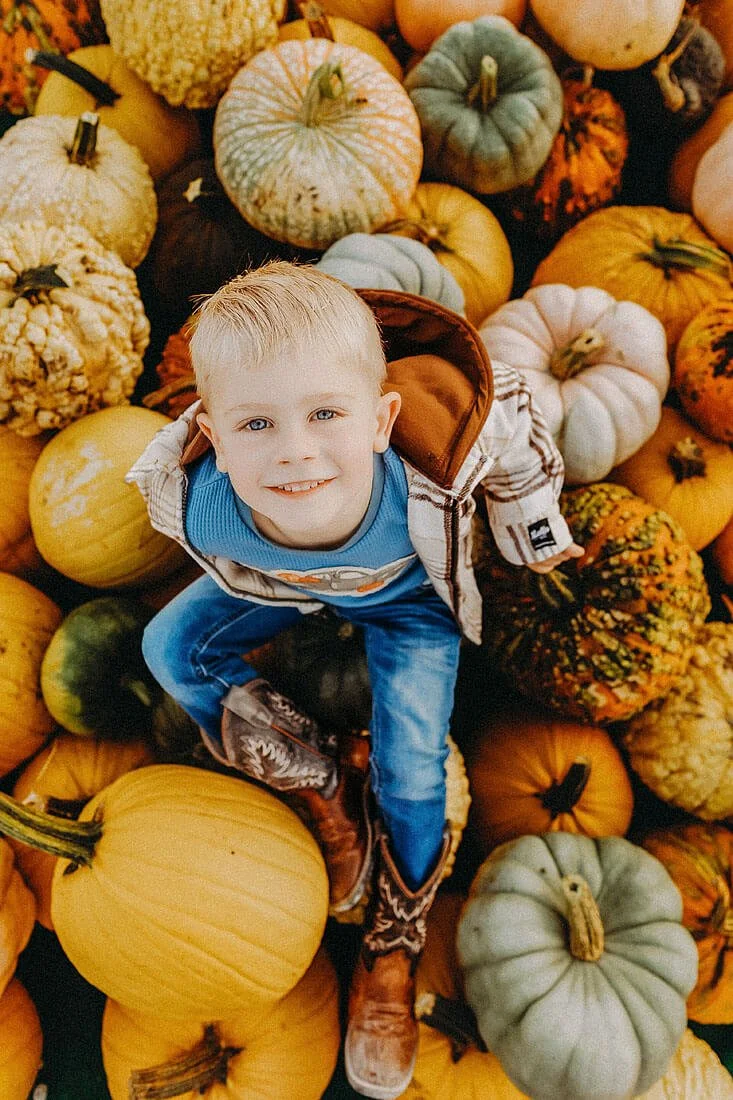 Young boy in cowboy boots smiles up at the camera while surrounded by colorful pumpkins and gourds at Dull’s Pumpkin Harvest