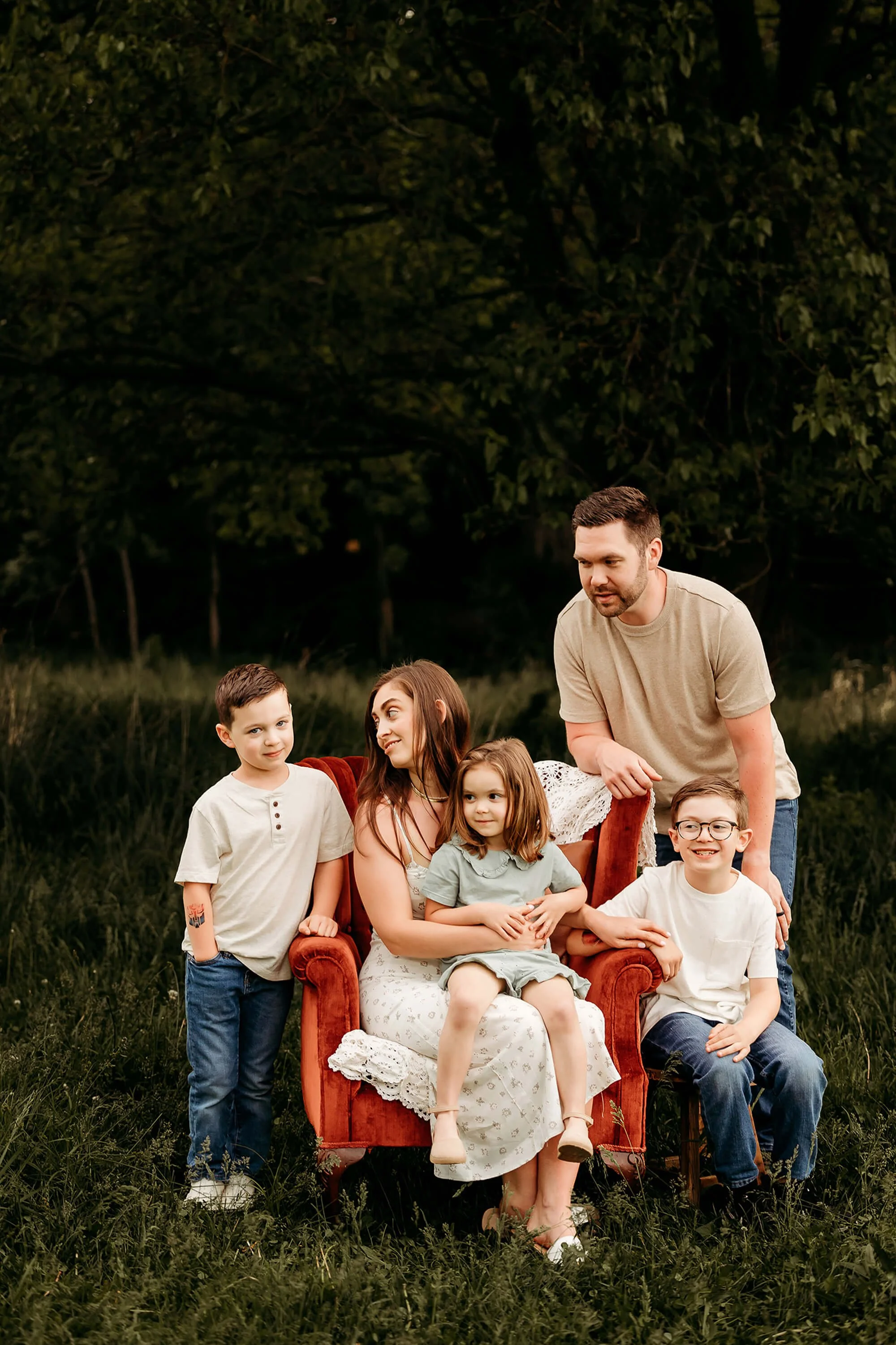 A family is seated on a red chair in a lush field, sharing a moment together in the open air.