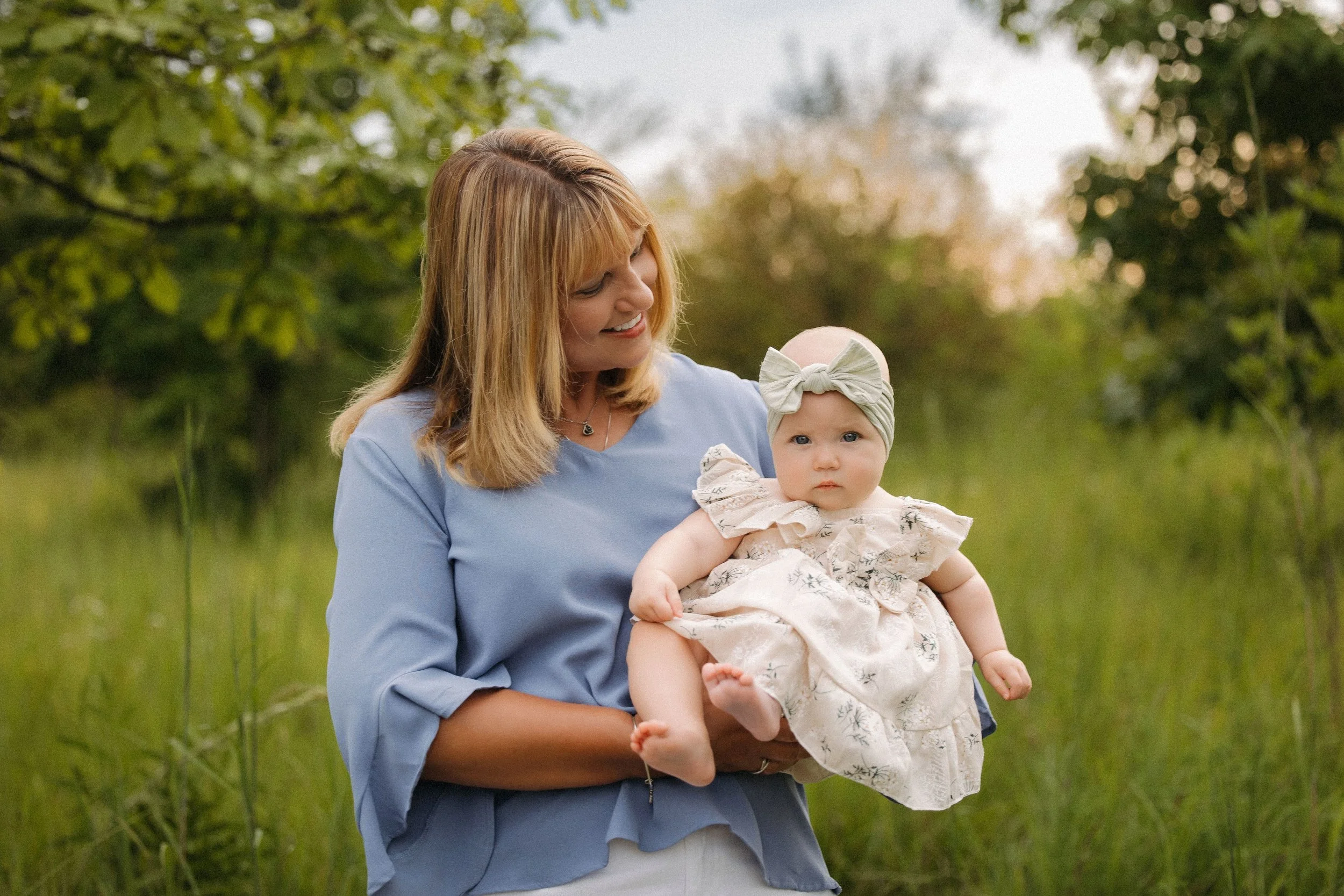 A woman cradles a baby in her arms while standing in a lush green field, surrounded by nature