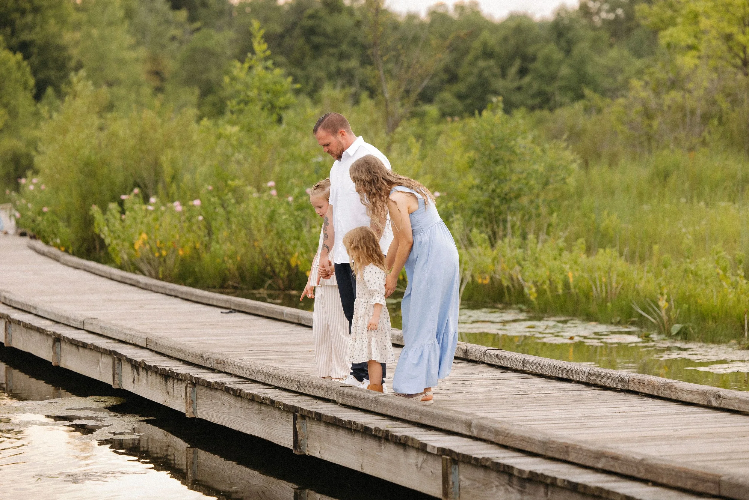 An extended family poses on a wooden bridge by a tranquil pond, surrounded by nature on a bright day