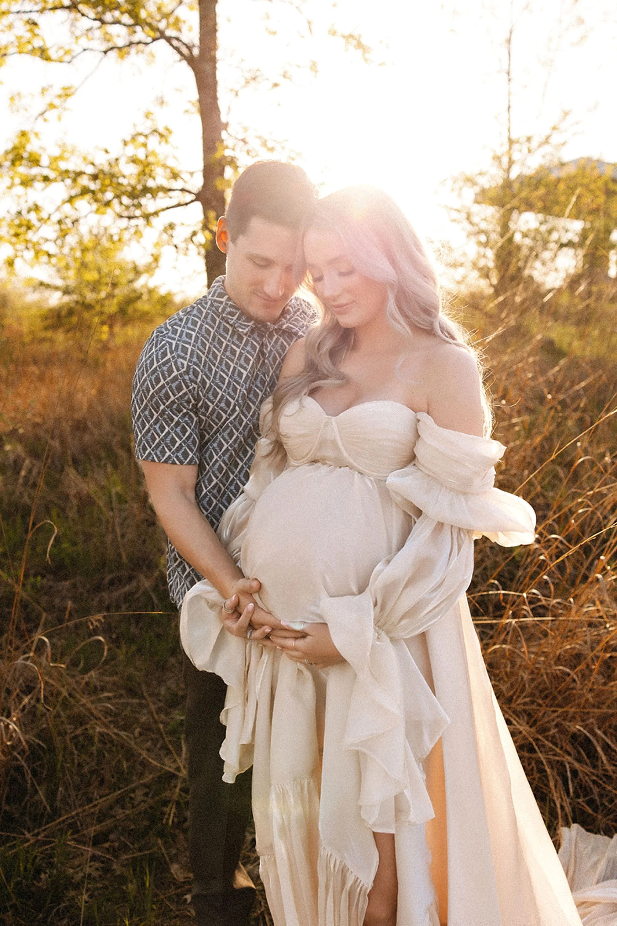 Maternity photos in Central Indiana featuring an expecting couple embracing outdoors at golden hour, husband holding his wife's baby bump in a sunlit field