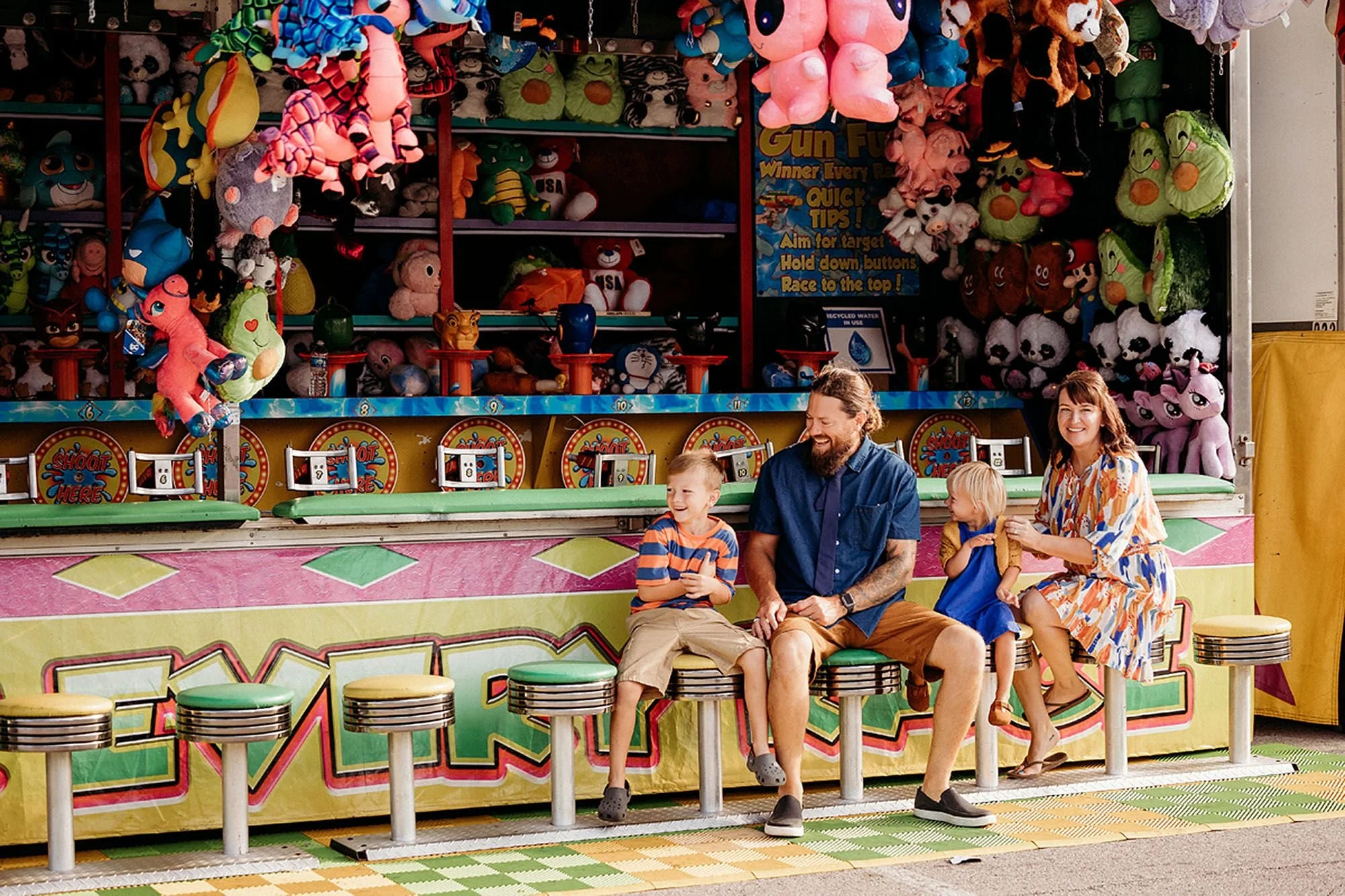 A family enjoys time together on a bench at a carnival, with bright lights and attractions in the background