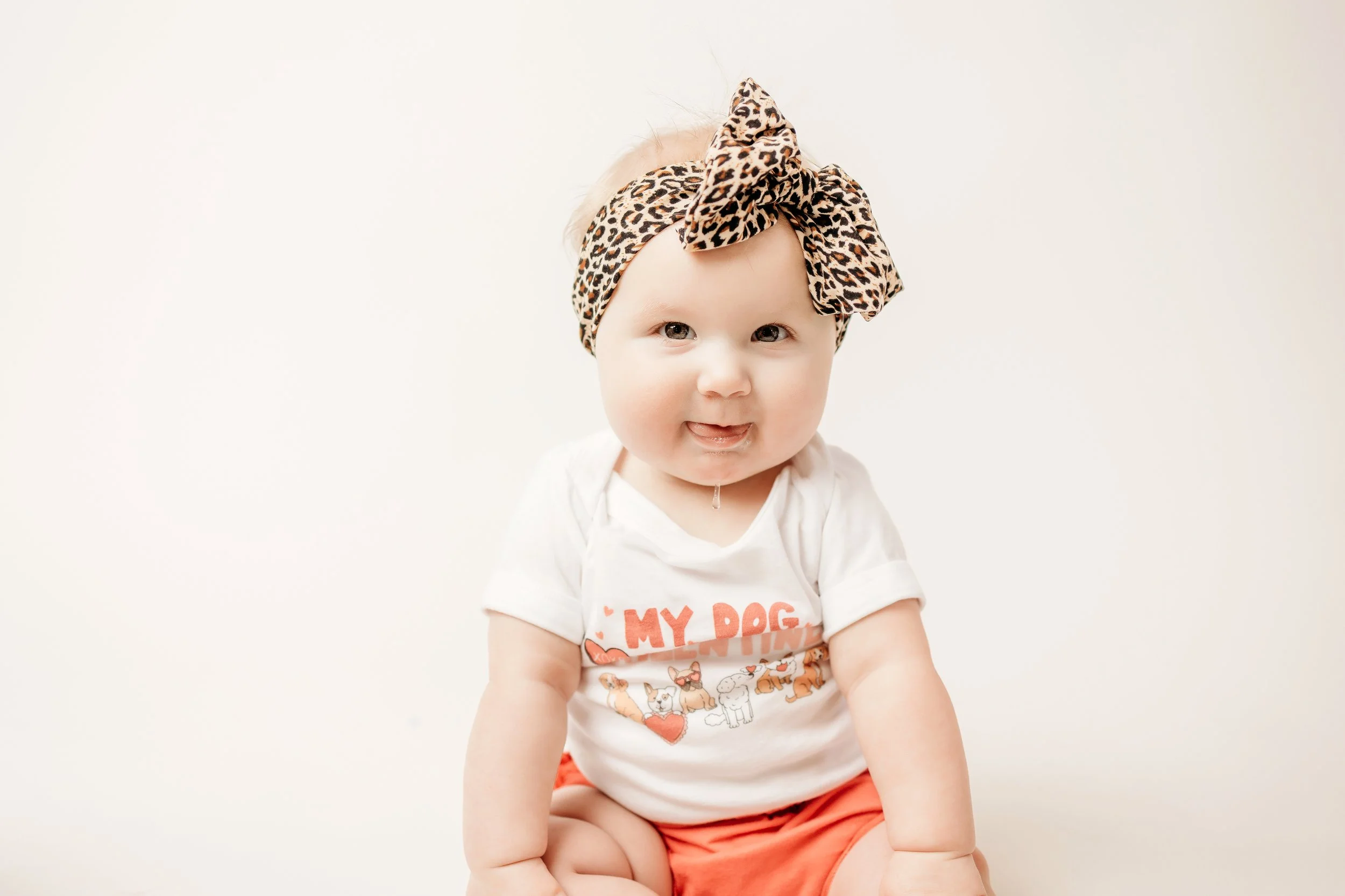 A baby girl dressed in a white shirt and a leopard print headband looks cheerful