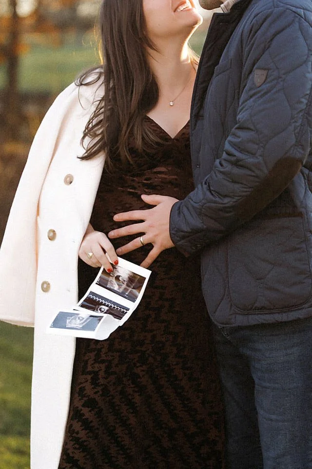 A heartfelt pregnancy announcement portrait as the couple gently cradles the baby bump, holding ultrasound photos against a backdrop of glowing autumn colors and soft golden-hour light.