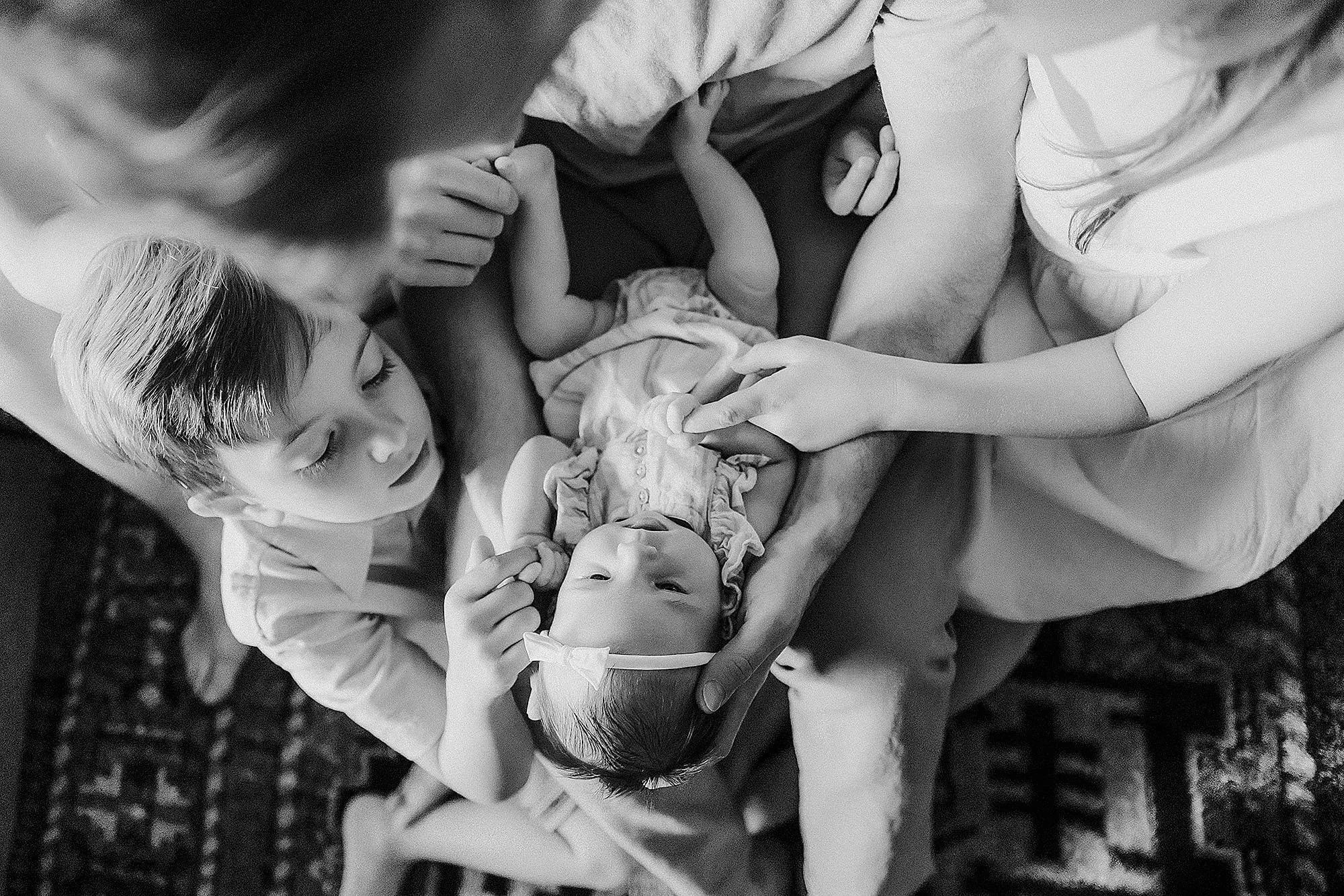 A family of four or five people lying on the floor, gathered around their sleeping infant. The children appear to be gently touching or holding the baby, showing affection and care.
