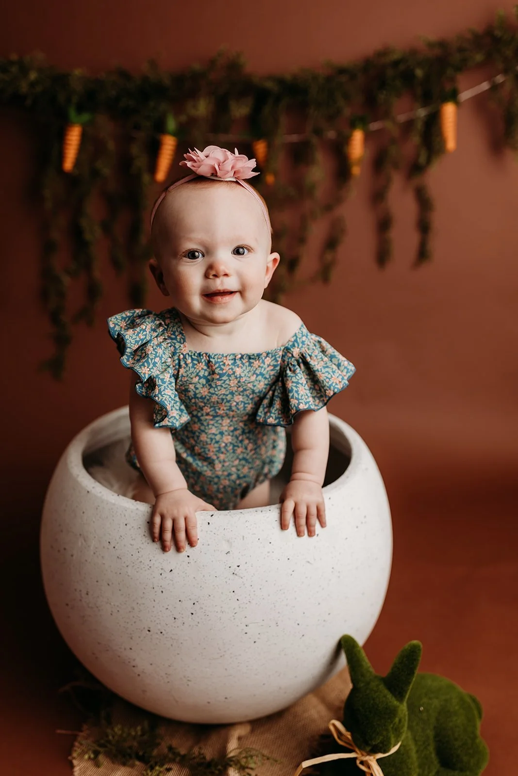 A smiling baby girl with a pink flower headband in a floral dress, leaning on a white speckled ceramic bowl, with a green stuffed bunny and a brown cloth in front, against a warm brown background with hanging green foliage and orange carrots.