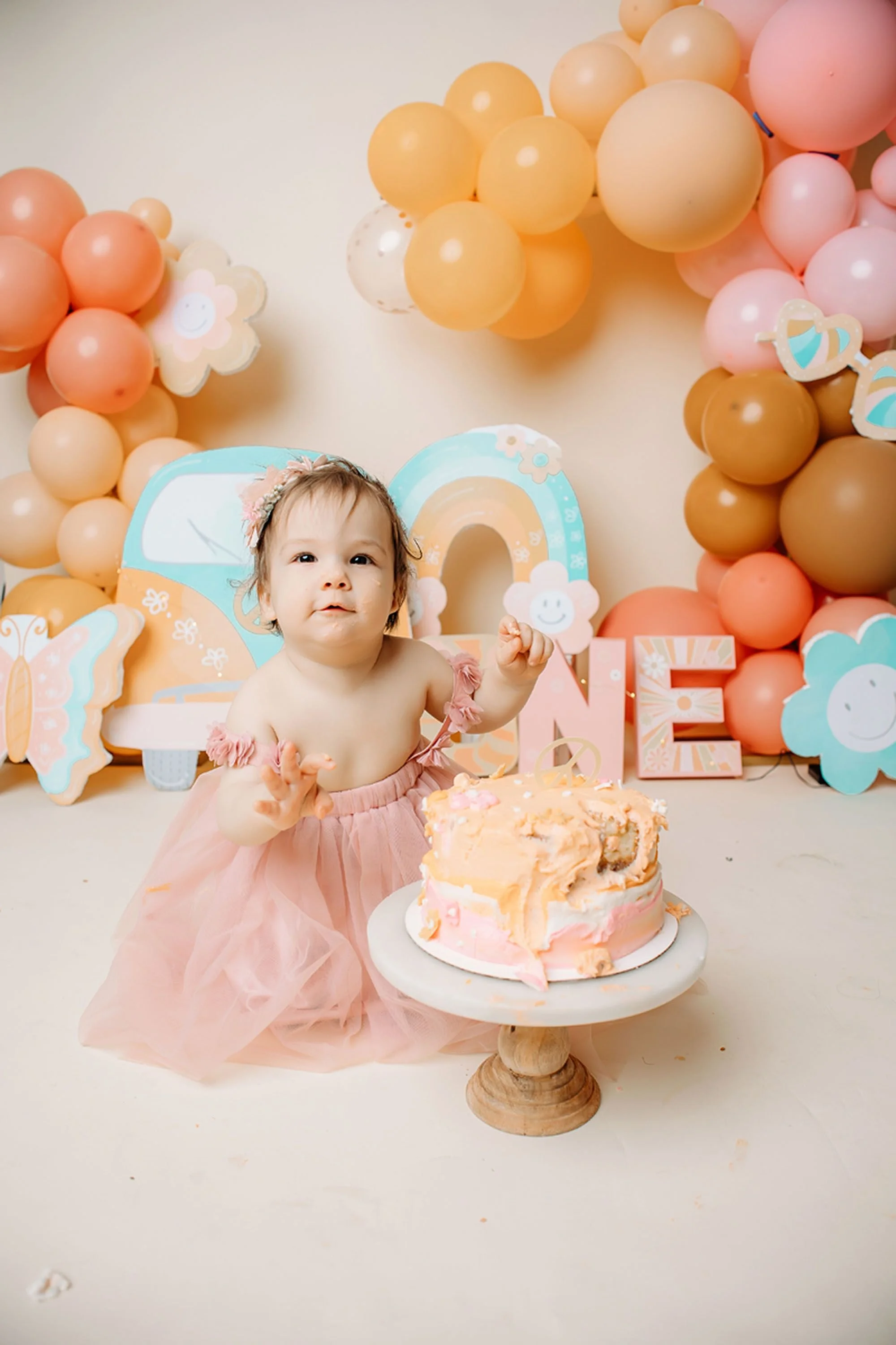 A happy baby celebrates her first birthday beside a pastel cake in a groovy one cake smash setup with warm orange balloons, retro rainbows, and floral decorations.