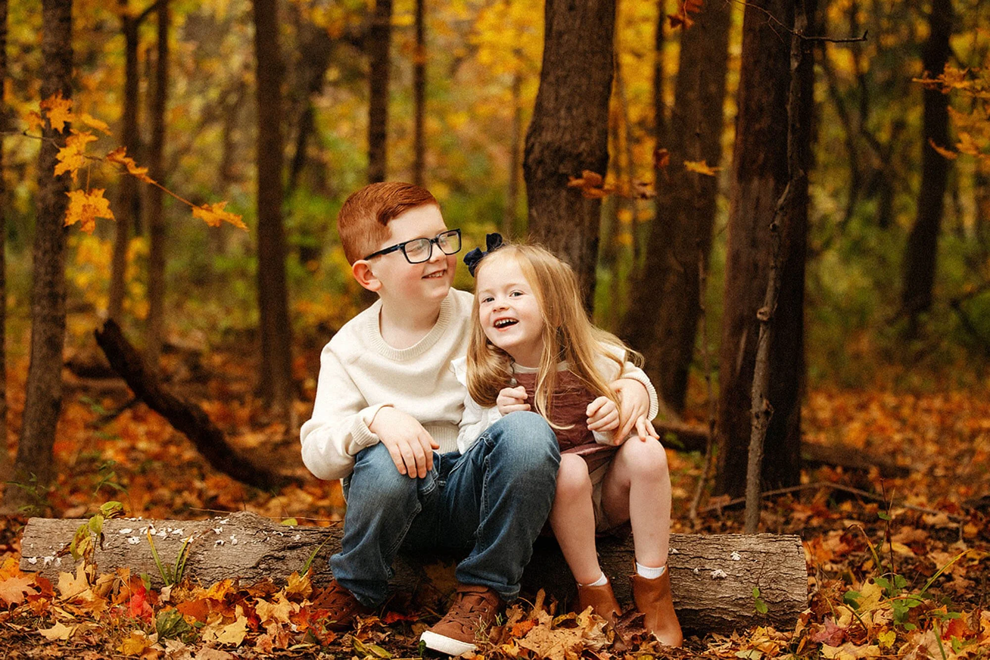 Family photographer capturing joyful sibling moments together in a beautiful autumn forest, surrounded by colorful fall leaves and warm natural light.