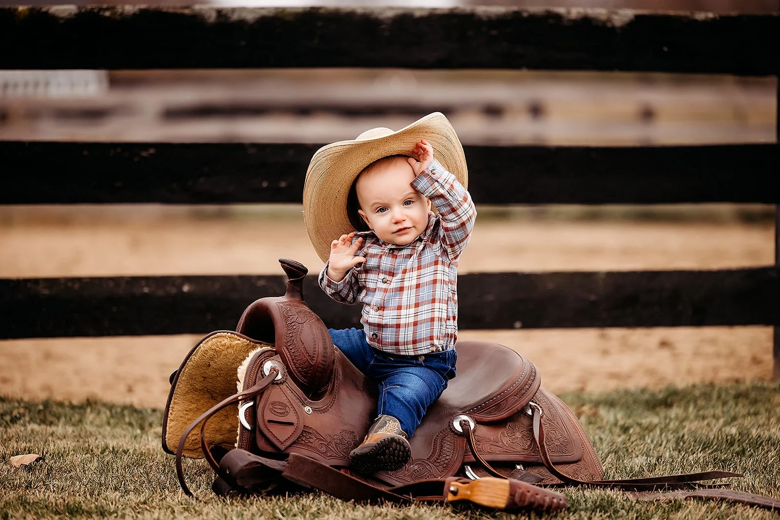 Young child sitting on a saddle with a cowboy hat, holding the hat with one hand, in an outdoor setting with grass and a black wooden fence in the background.