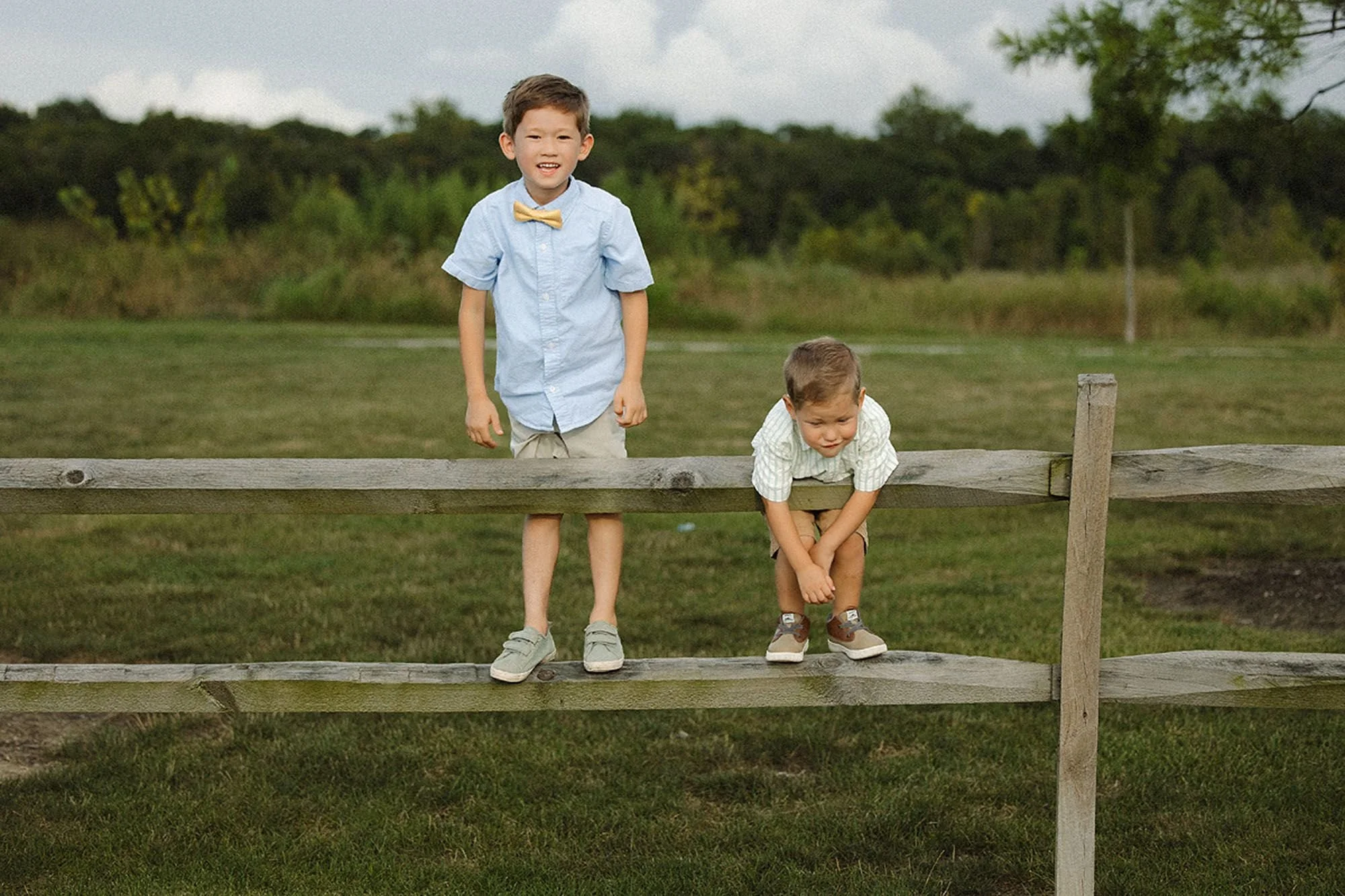 Two young brothers standing and sitting on a wooden split-rail fence in a green field during a Flat Fork Creek Park family photo session