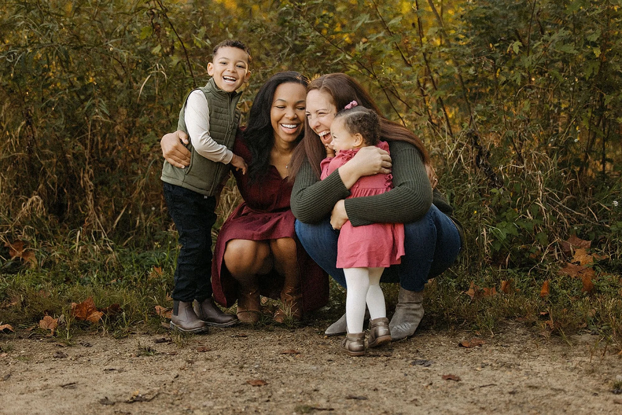 Family of four laughing and hugging on a wooded path, both mothers crouched between their kids at Virginia B. Fairbanks Art & Nature Park, family photos capturing a joyful full-family embrace