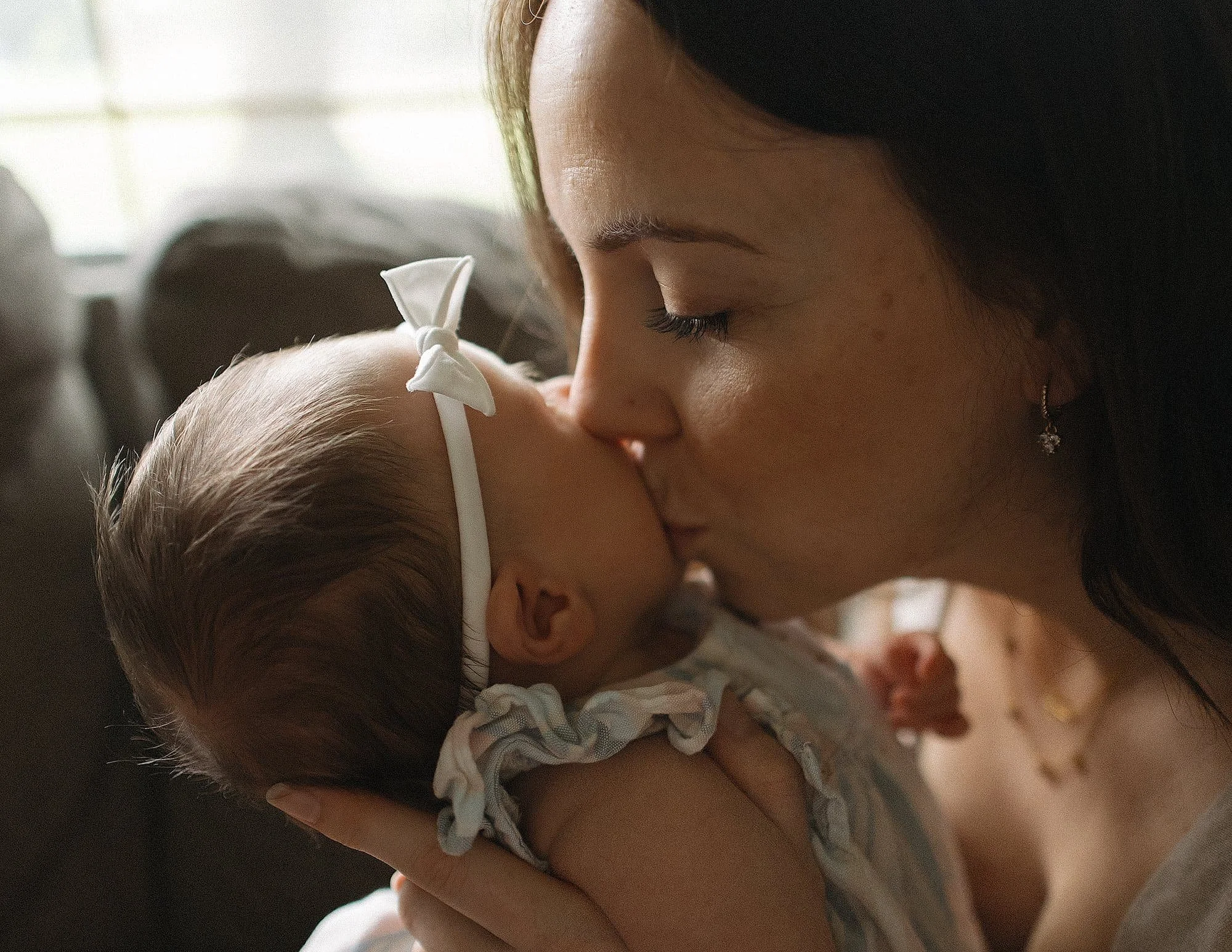 A woman gently kisses a baby on the forehead indoors with natural light.
