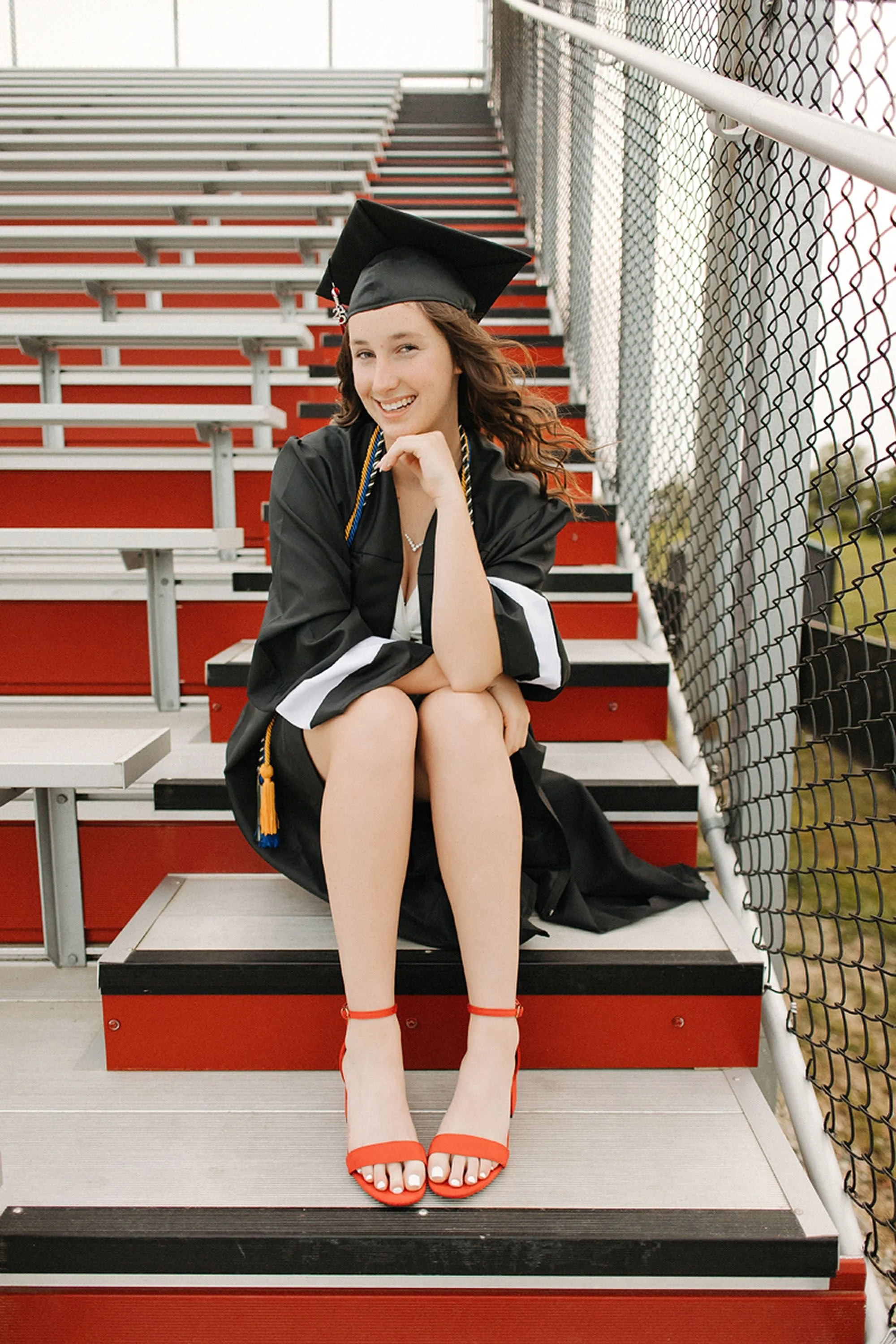 Smiling graduate sitting on stadium bleachers in a black cap and gown with bright red heels during Sheridan High School Senior Photos.