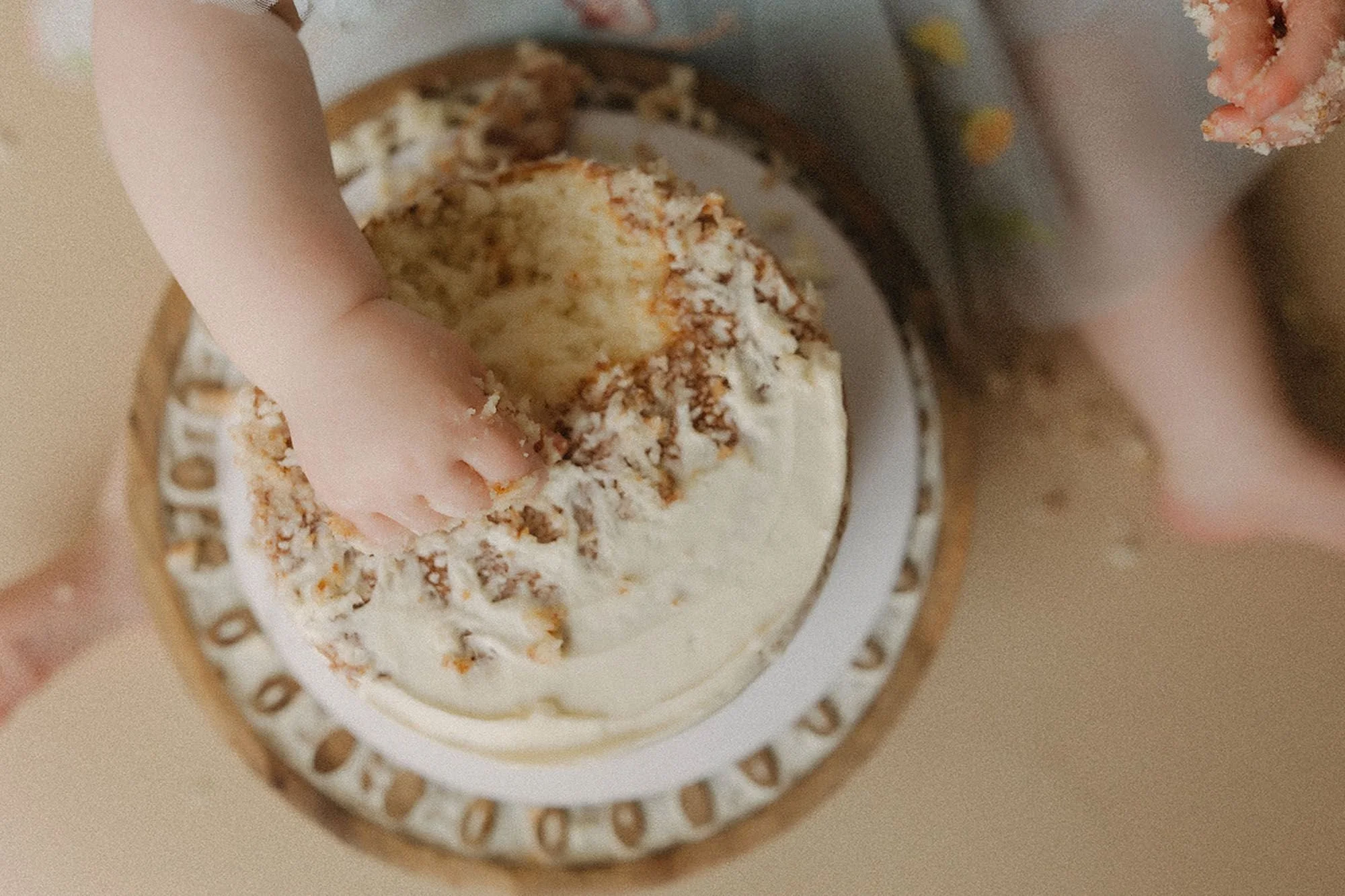 Close-up overhead shot of a baby's hand digging into a white buttercream cake with golden coconut coating, simple cake smash detail photography on a decorative wooden cake stand
