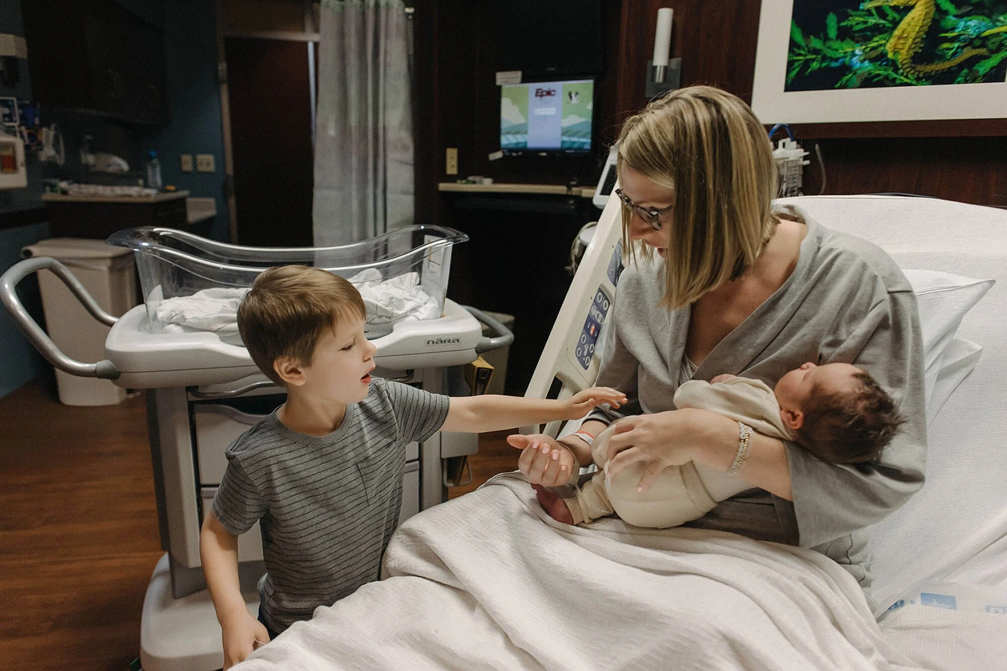 Mother holds her newborn in a hospital bed, with two children nearby, highlighting a joyful Fresh 48 session during a newborn session