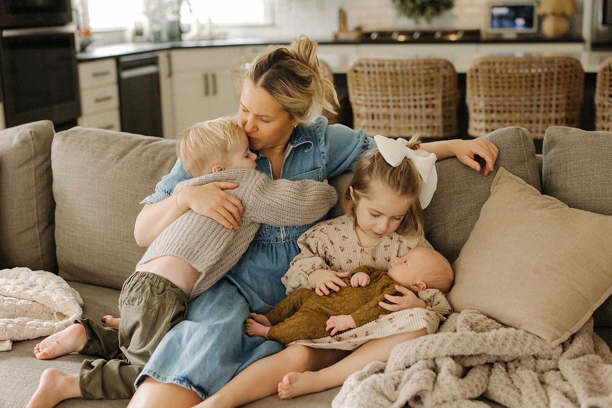 Heartwarming in-home newborn family photography featuring a loving mother cuddling her older son while a proud sister gently holds their newborn baby on a cozy living room couch