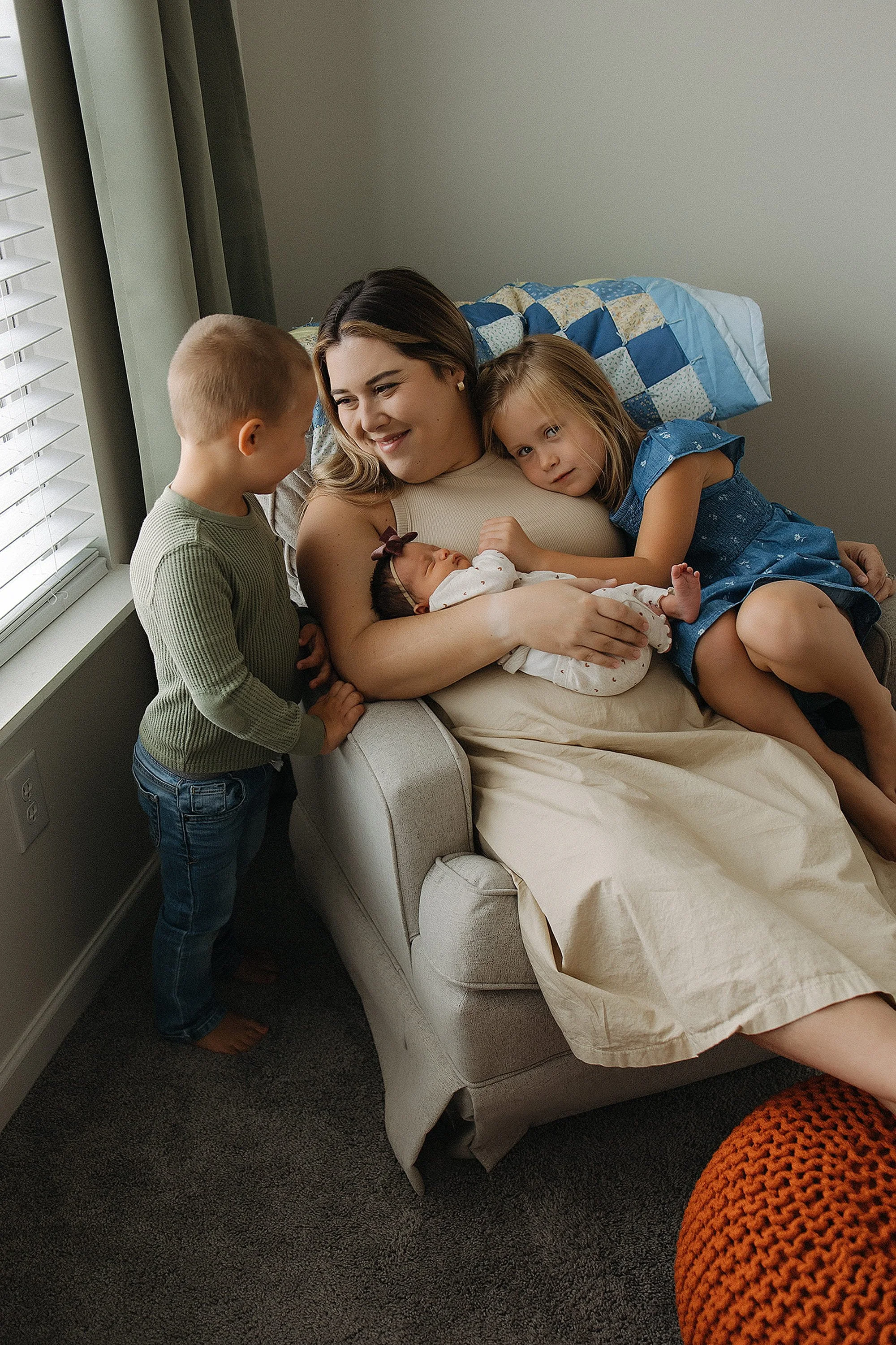 A woman sitting on a couch with two young girls and a newborn baby. The woman is holding the baby, while the older girl rests her head on the woman's shoulder. The younger boy stands beside the couch, looking at the woman and baby. The scene takes pl