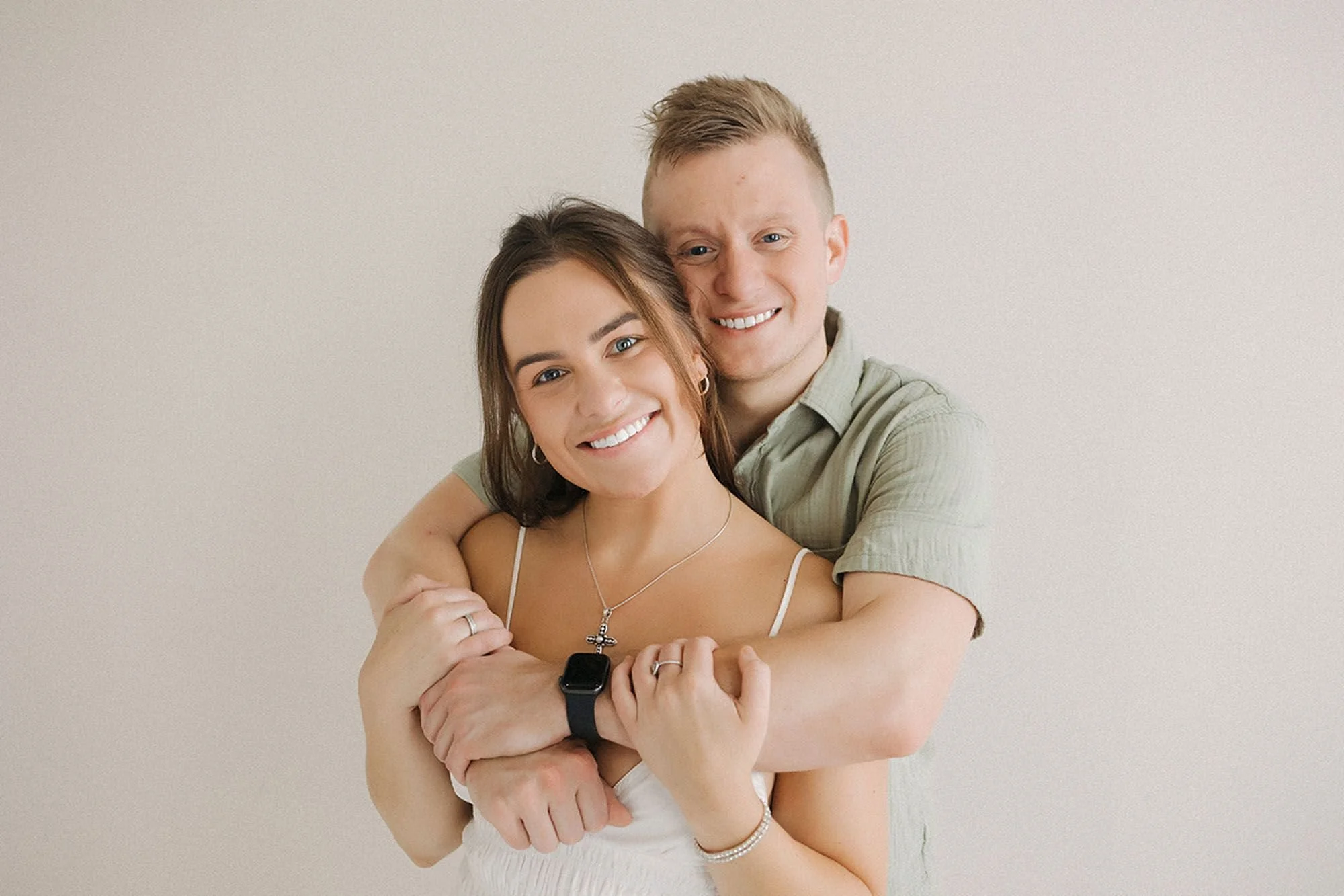 Smiling couple posing for a studio portrait. He stands behind her with his arms wrapped around her shoulders while she holds his forearms, both laughing warmly against a soft neutral backdrop