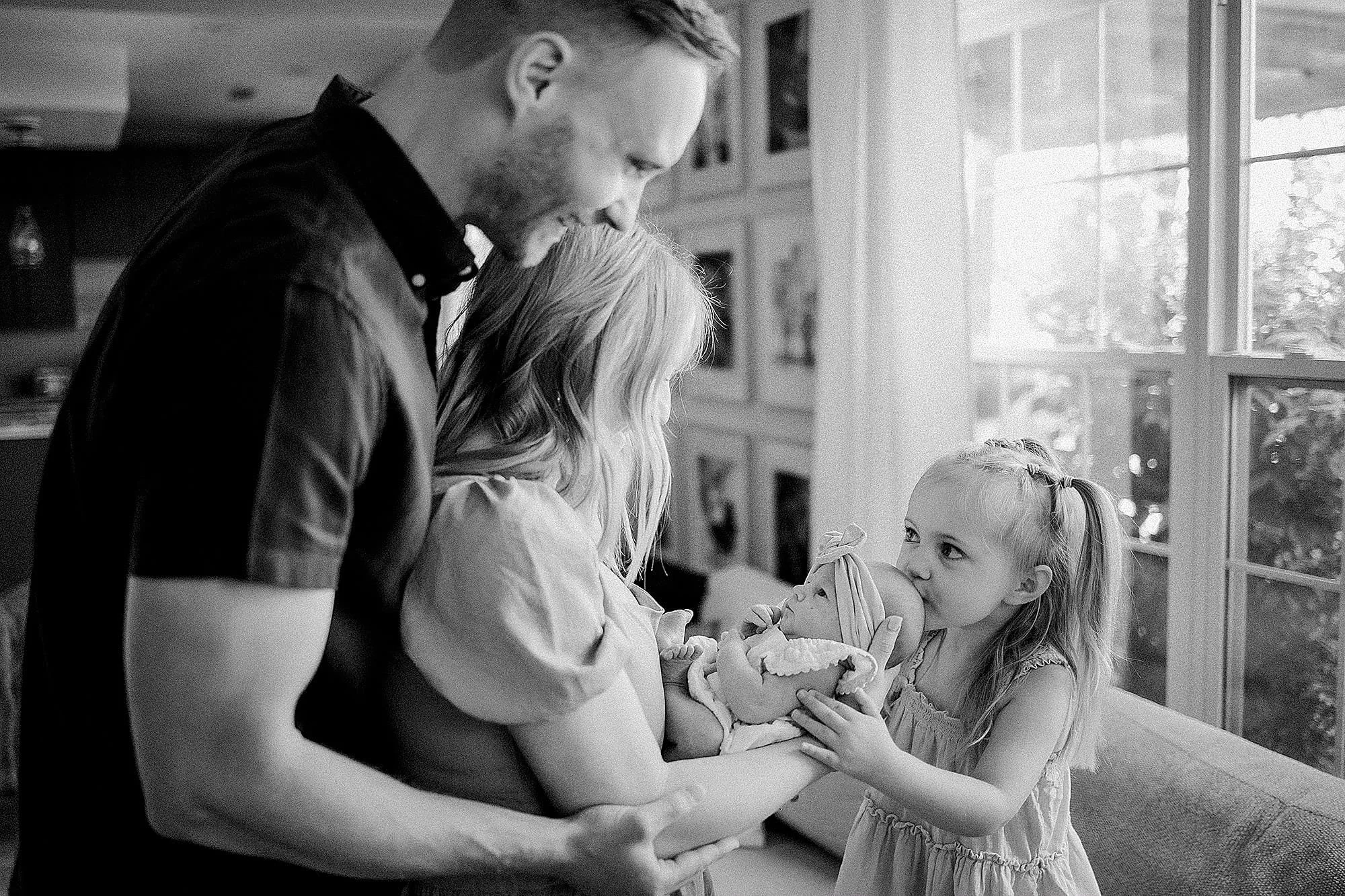 A family with young children by a window, with a woman holding a newborn baby, a girl kissing the baby, and a man leaning over them, all smiling.