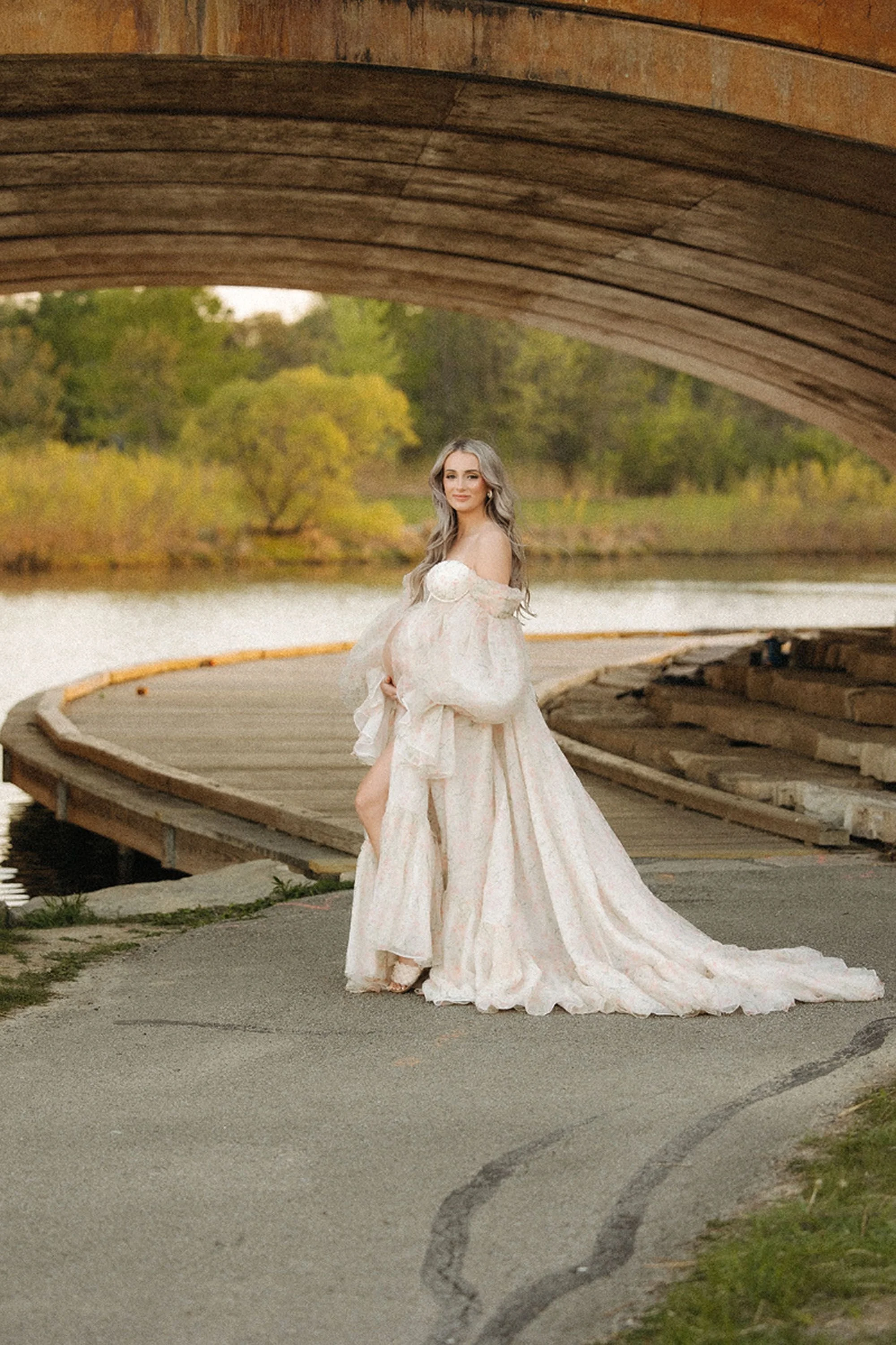 Maternity photos in Central Indiana showing a pregnant woman in a flowing cream gown standing on a curved boardwalk beneath a wooden bridge by the lake