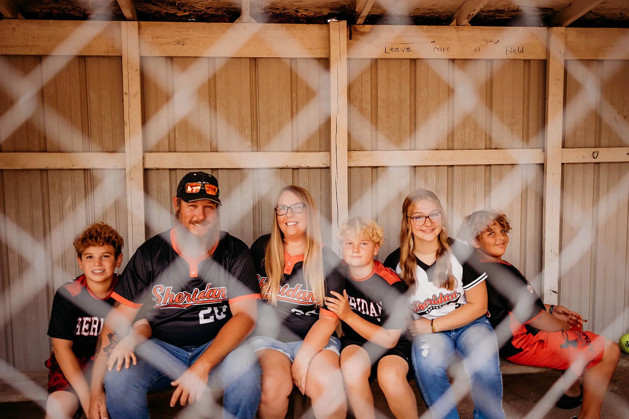 A family sits together in a dugout behind a fence, smiling during a pregnancy announcement with their older kids, captured by The Heart Narrative, an Indiana family photographer.