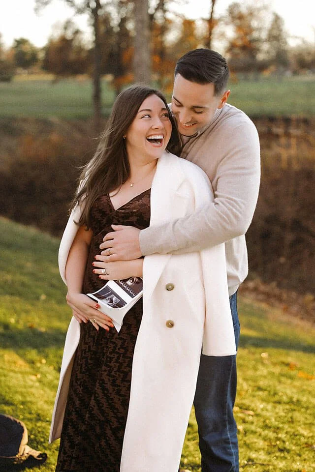 Pregnant woman laughs with her partner as they celebrate their growing family, surrounded by autumn scenery and beautiful natural golden light.