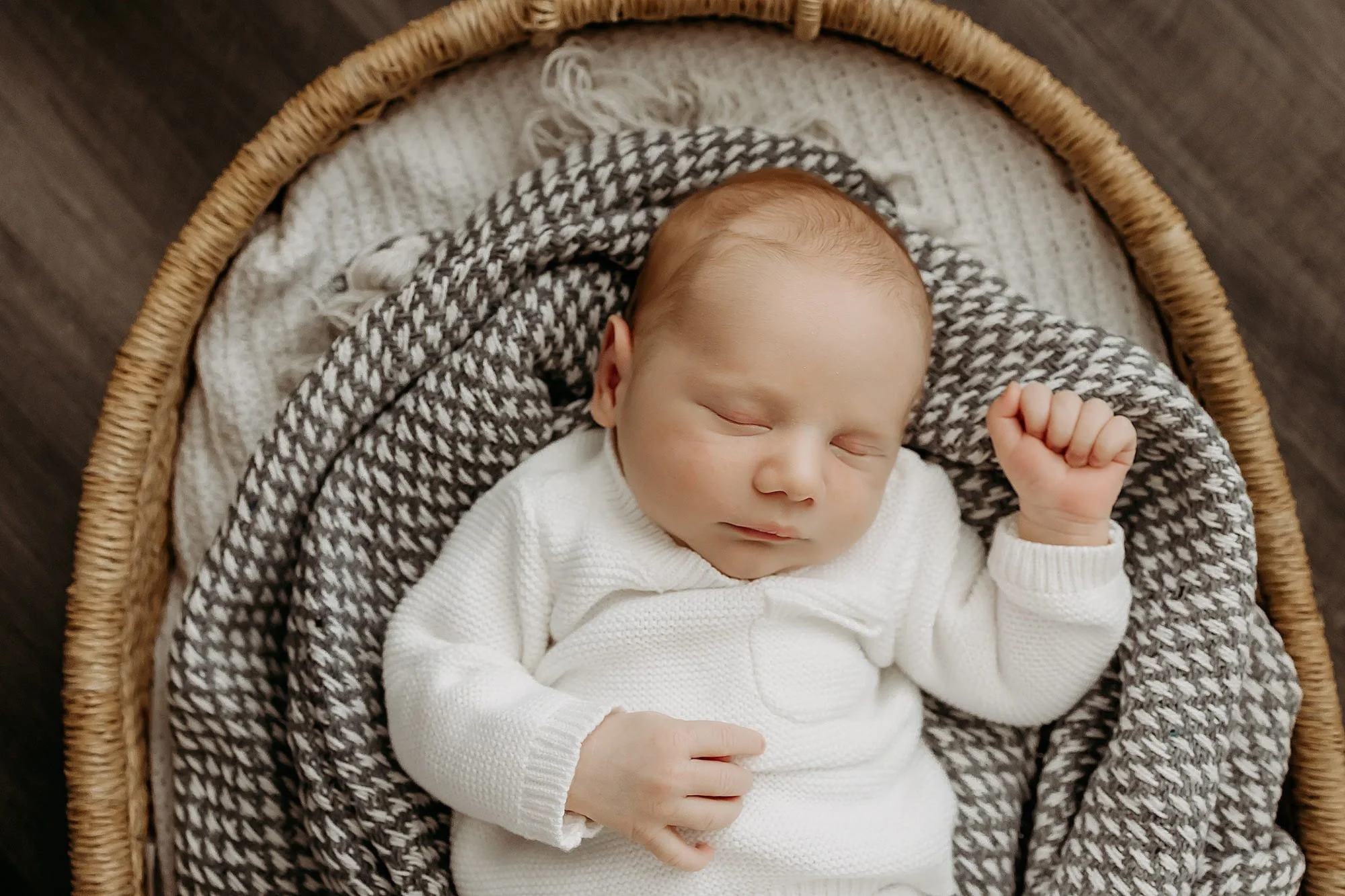 A sleeping baby in a white sweater lying in a woven bassinet with a patterned blanket.