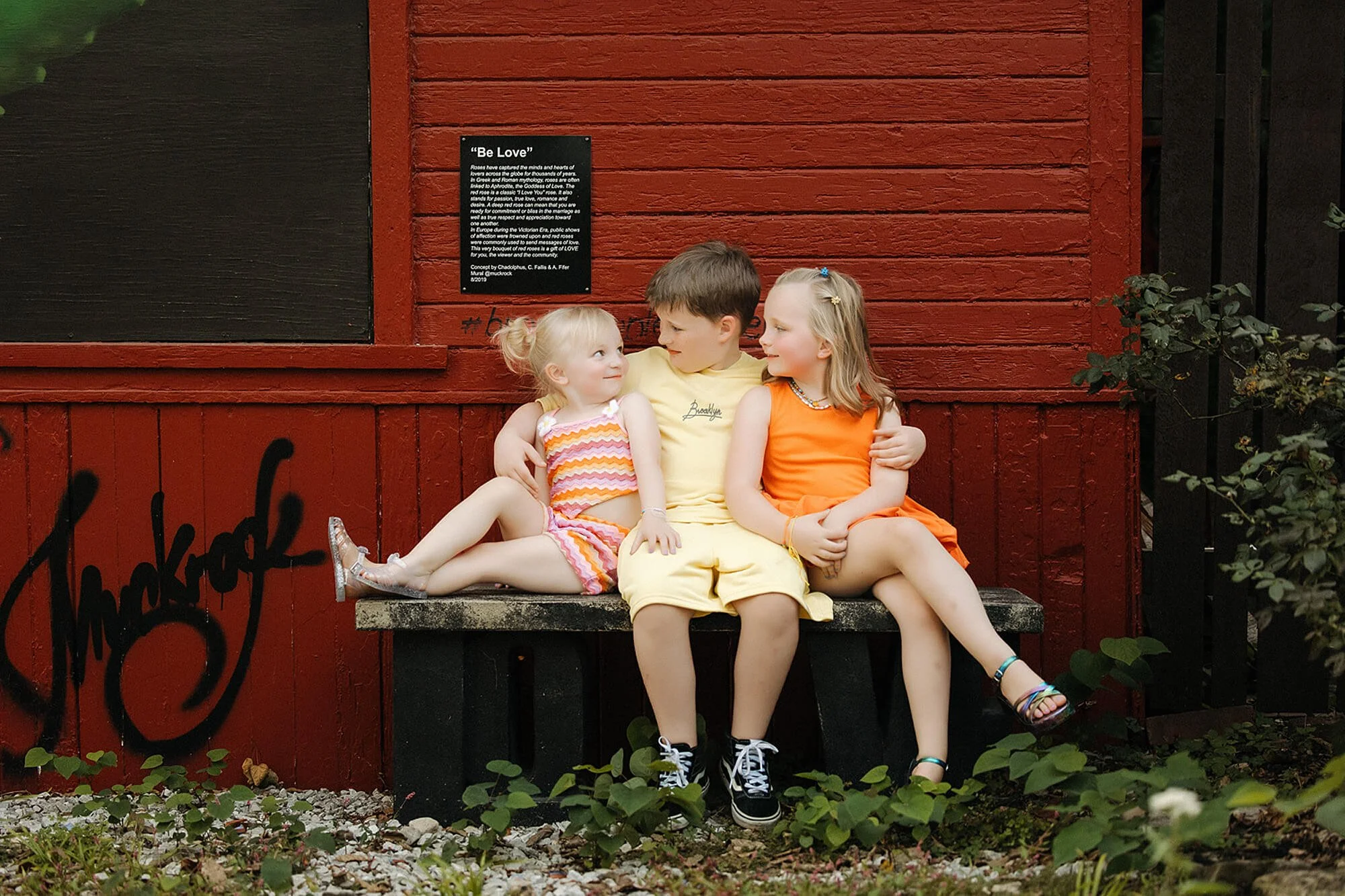 3 siblings sit on bench by red garage door in Flower Alley, Broadripple in Indianapolis during their fun and unique family photoshoot by Lauren Lewis