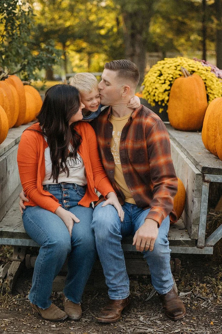 Parents cuddle their laughing young son while sitting on a wagon surrounded by pumpkins and fall flowers at Dull’s Pumpkin Harvest