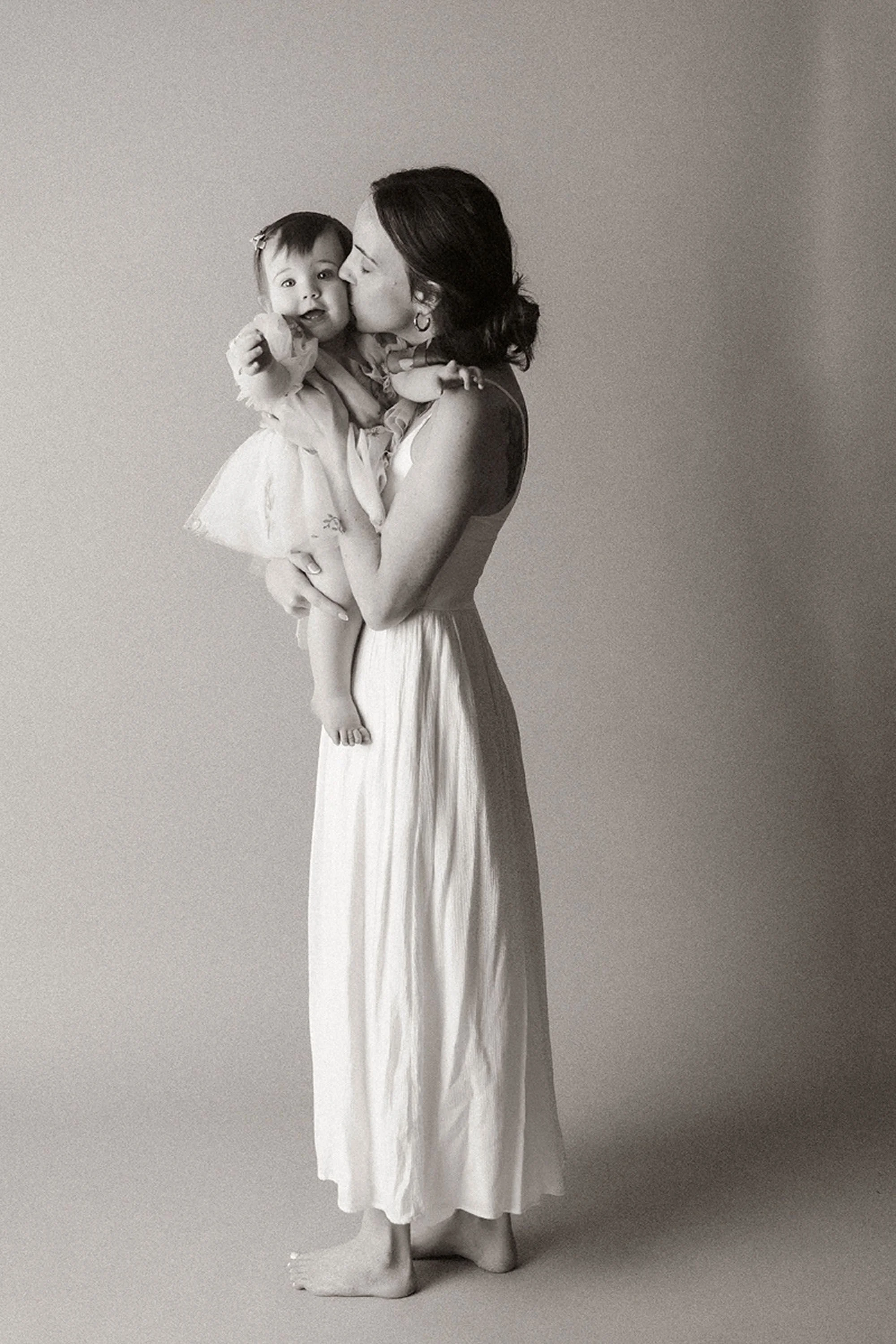 Simple Cake Smash Photos of a mother kissing her baby girl's cheek during a first birthday session, capturing a sweet and timeless moment in a minimalist studio with soft, natural styling.