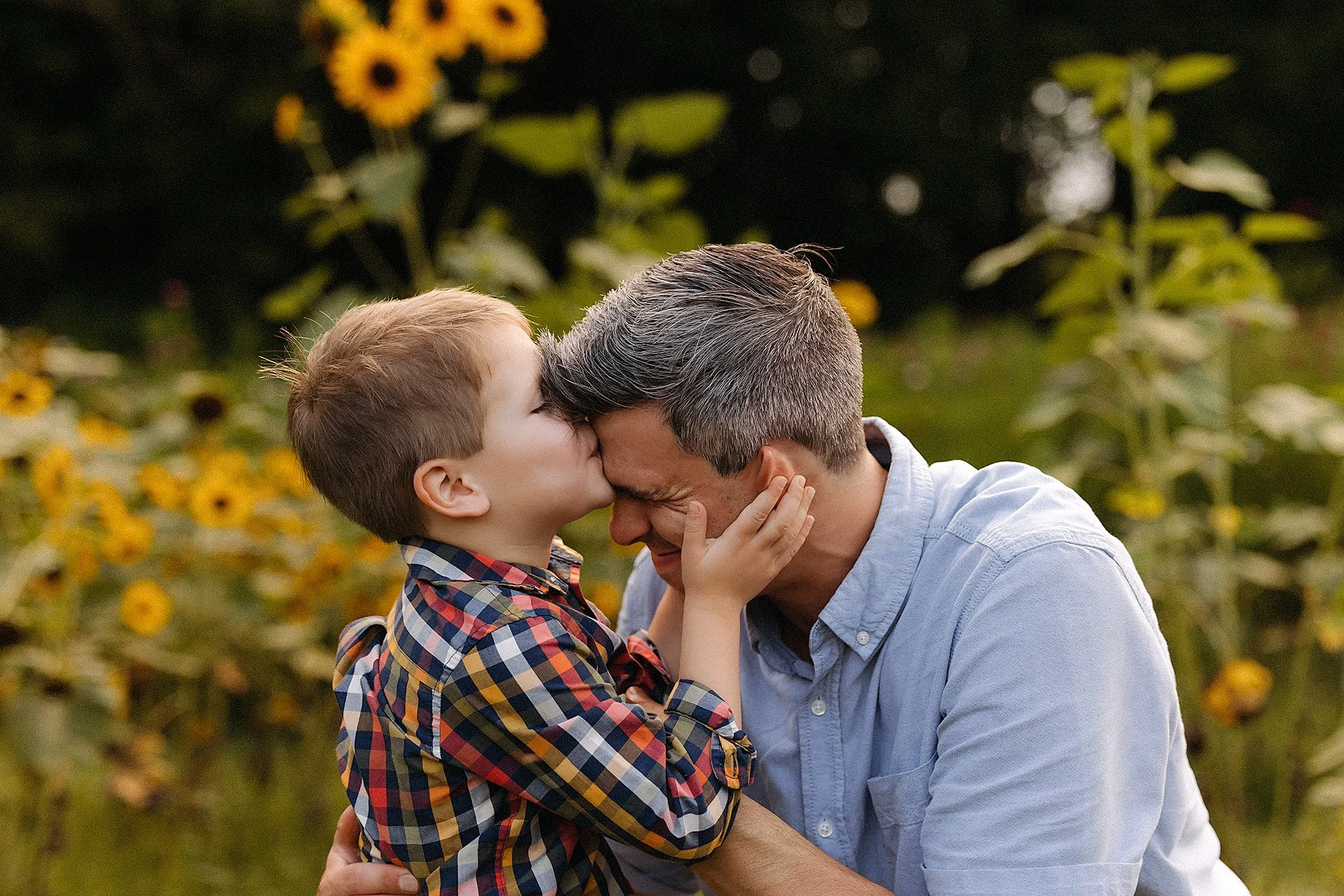 A man and a boy share a kiss in front of vibrant sunflowers during a Mini Session photography event.