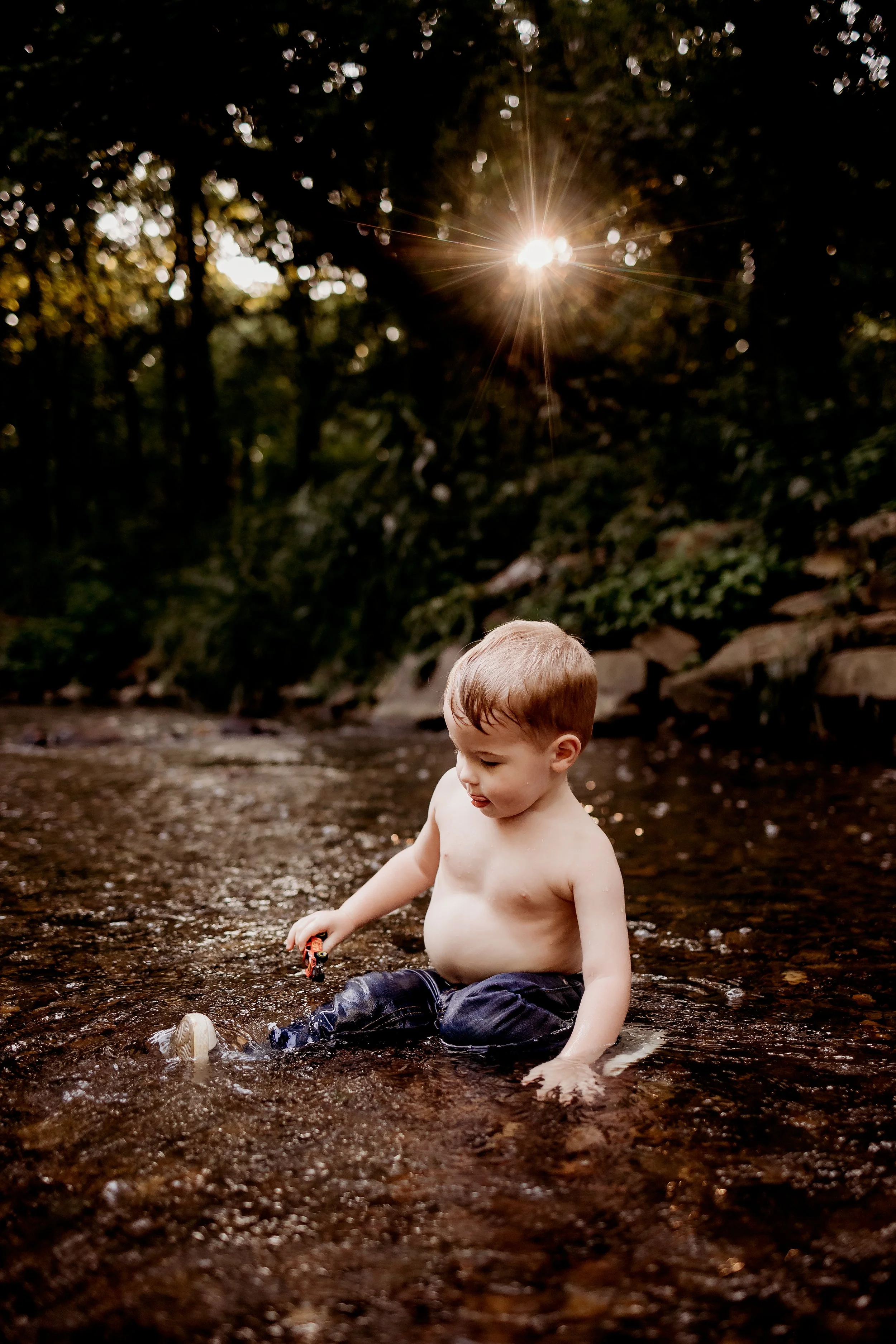 A young shirtless boy sitting in a shallow creek, playing with a toy car, surrounded by trees with sunlight shining through.