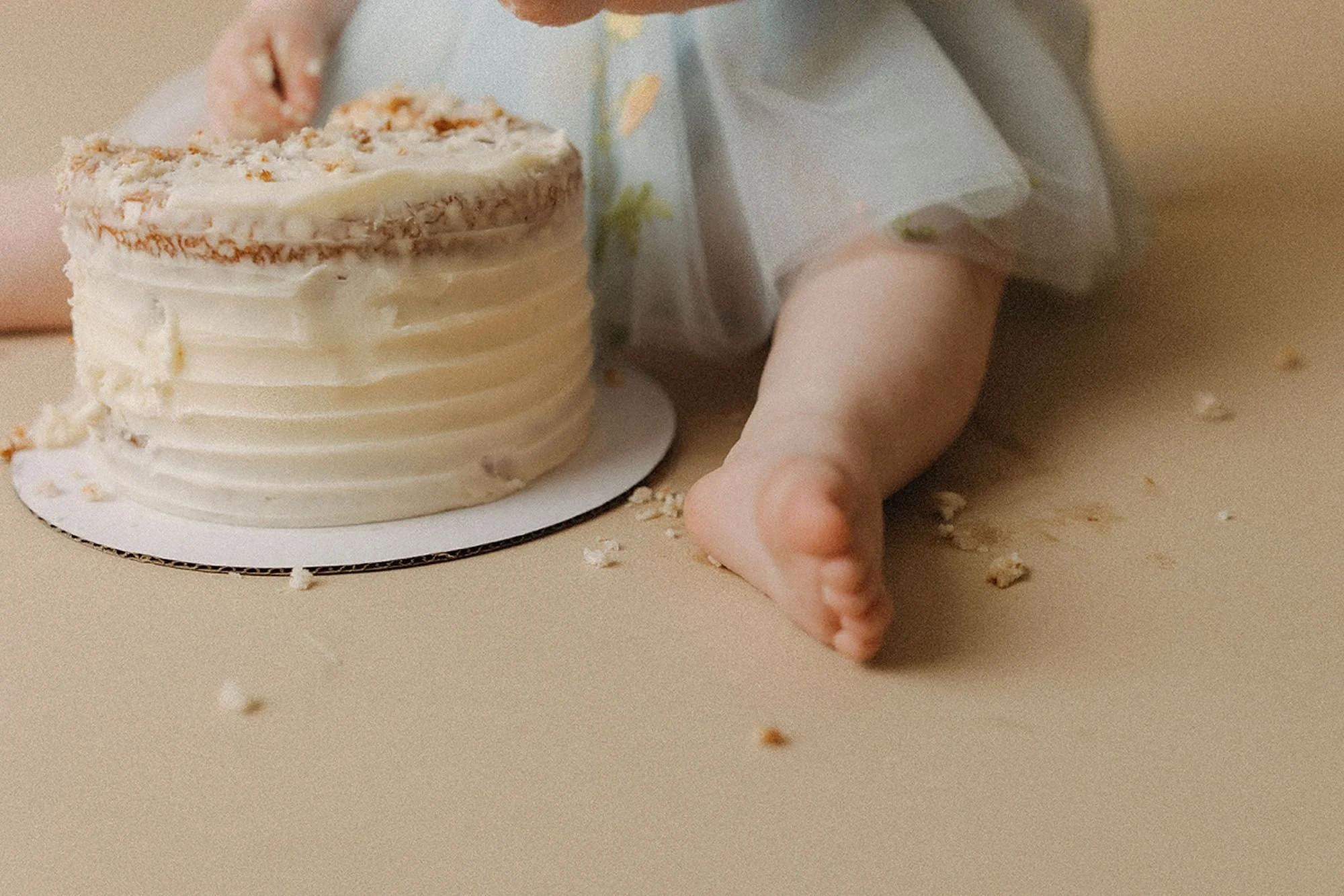 Close-up of a baby's bare feet next to a white buttercream cake with crumbs scattered around, simple cake smash photography detail shot