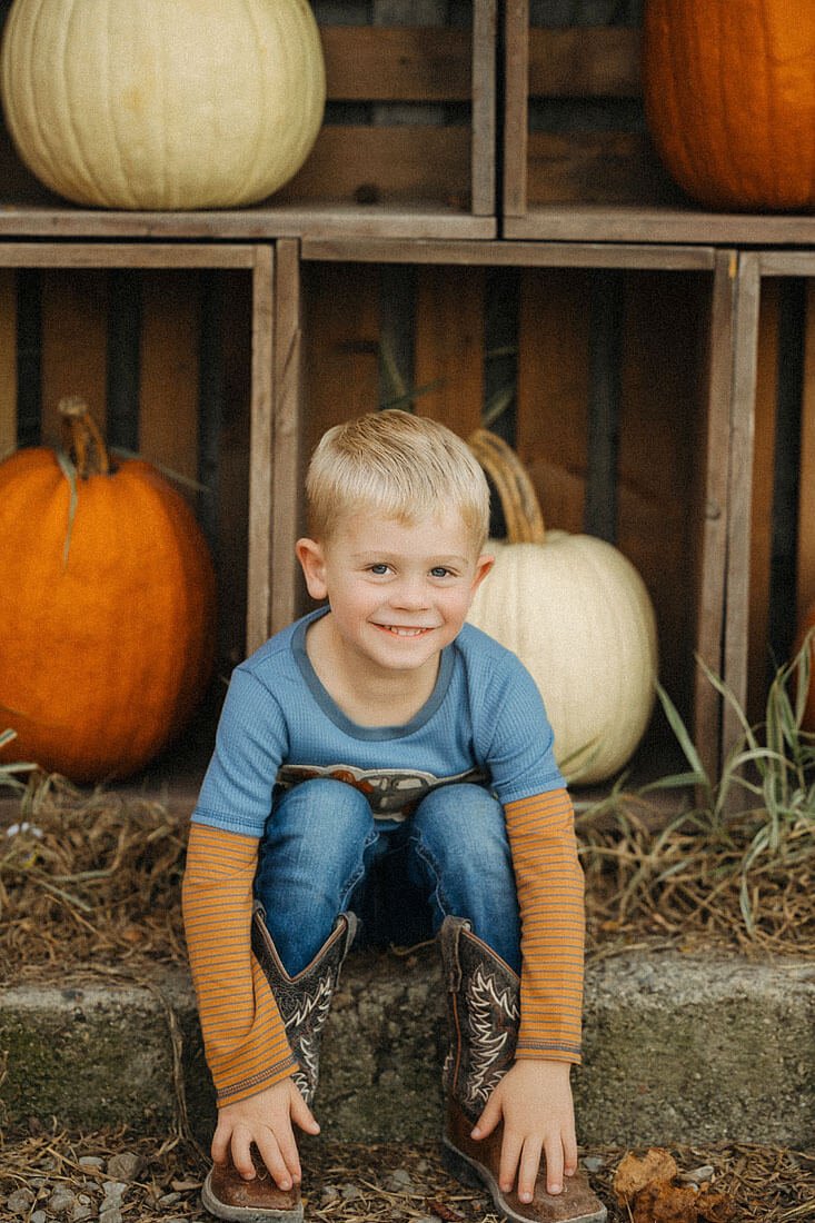 Smiling young boy wearing cowboy boots sits beside orange and white pumpkins during a relaxed fall family session at Dull’s Pumpkin Harvest