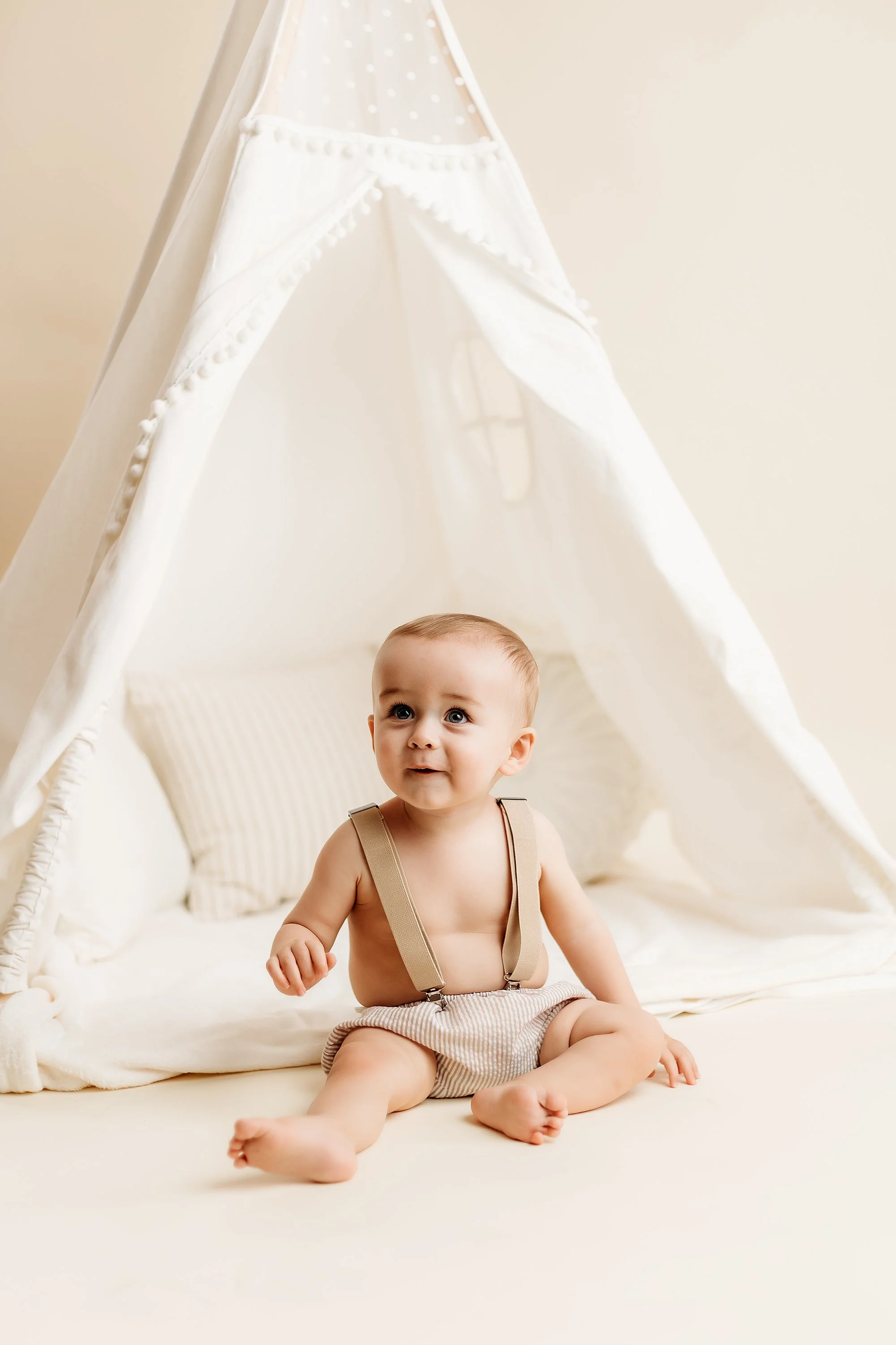 A baby sitting on a cream-colored blanket in front of a white fabric teepee tent with pillows.
