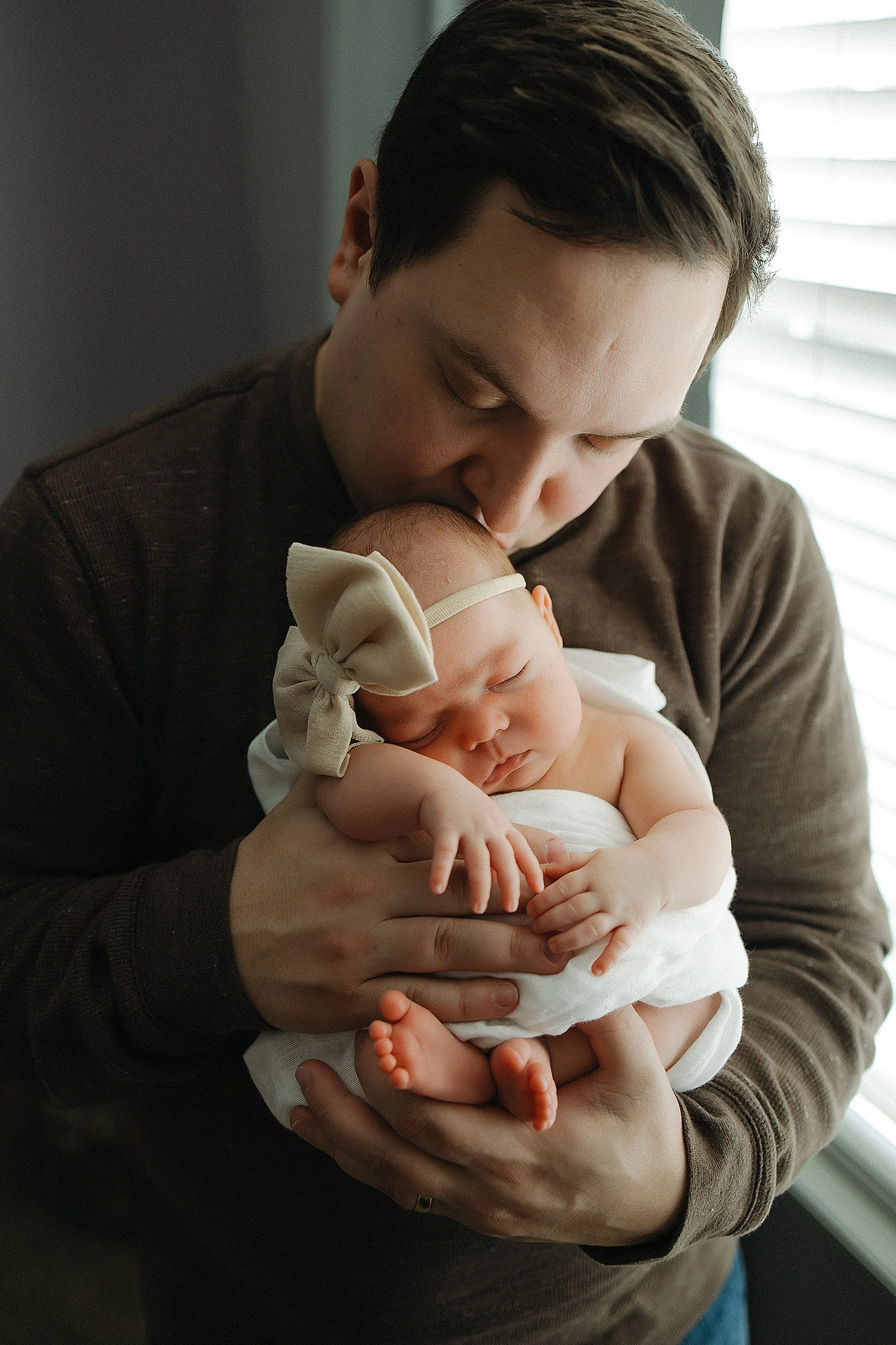 A man holding a sleeping baby girl, who is wearing a beige bow headband and white outfit, near a window with blinds.