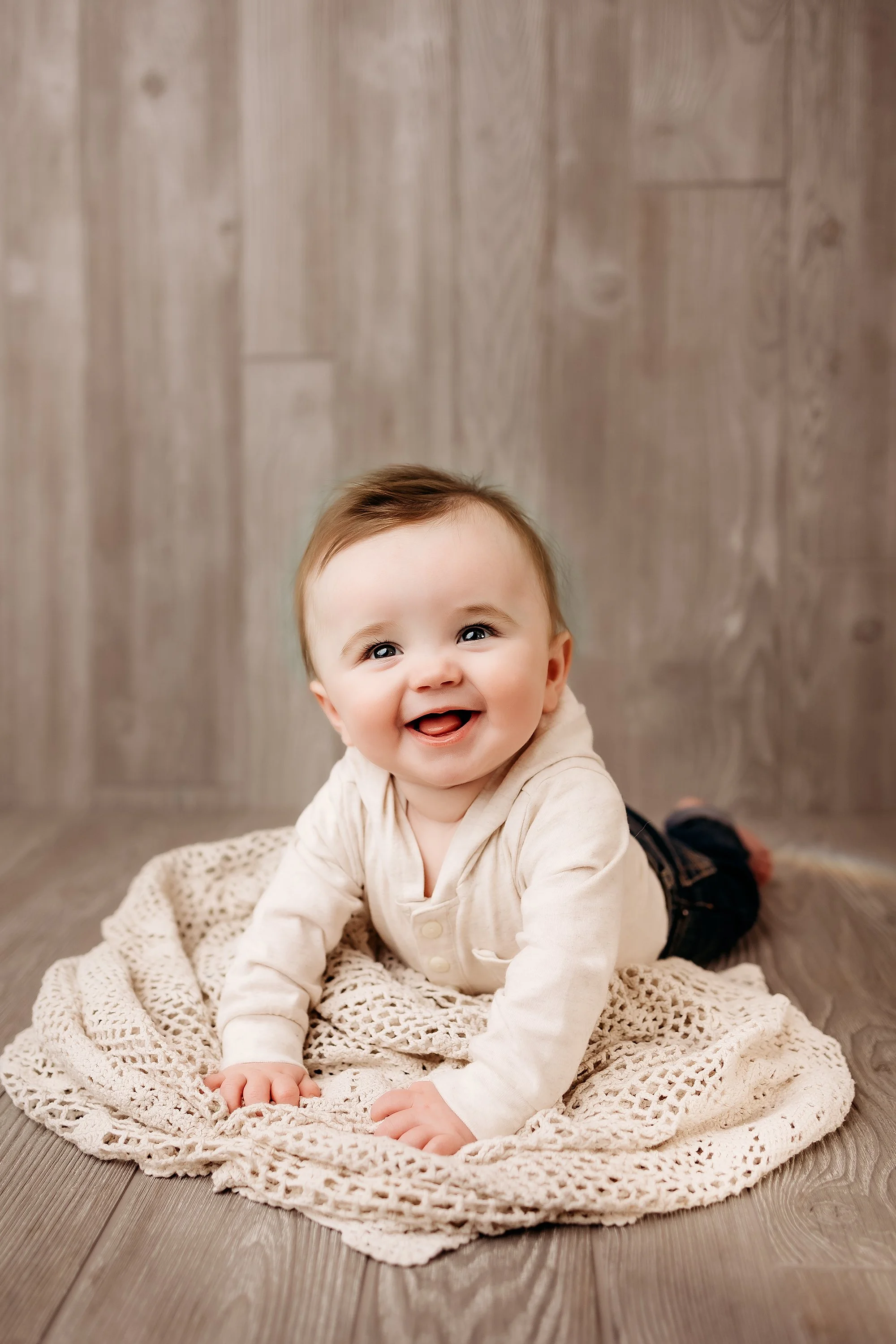 A cheerful baby with big eyes and a big smile laying on a crocheted blanket on a wooden floor with a wooden wall background.