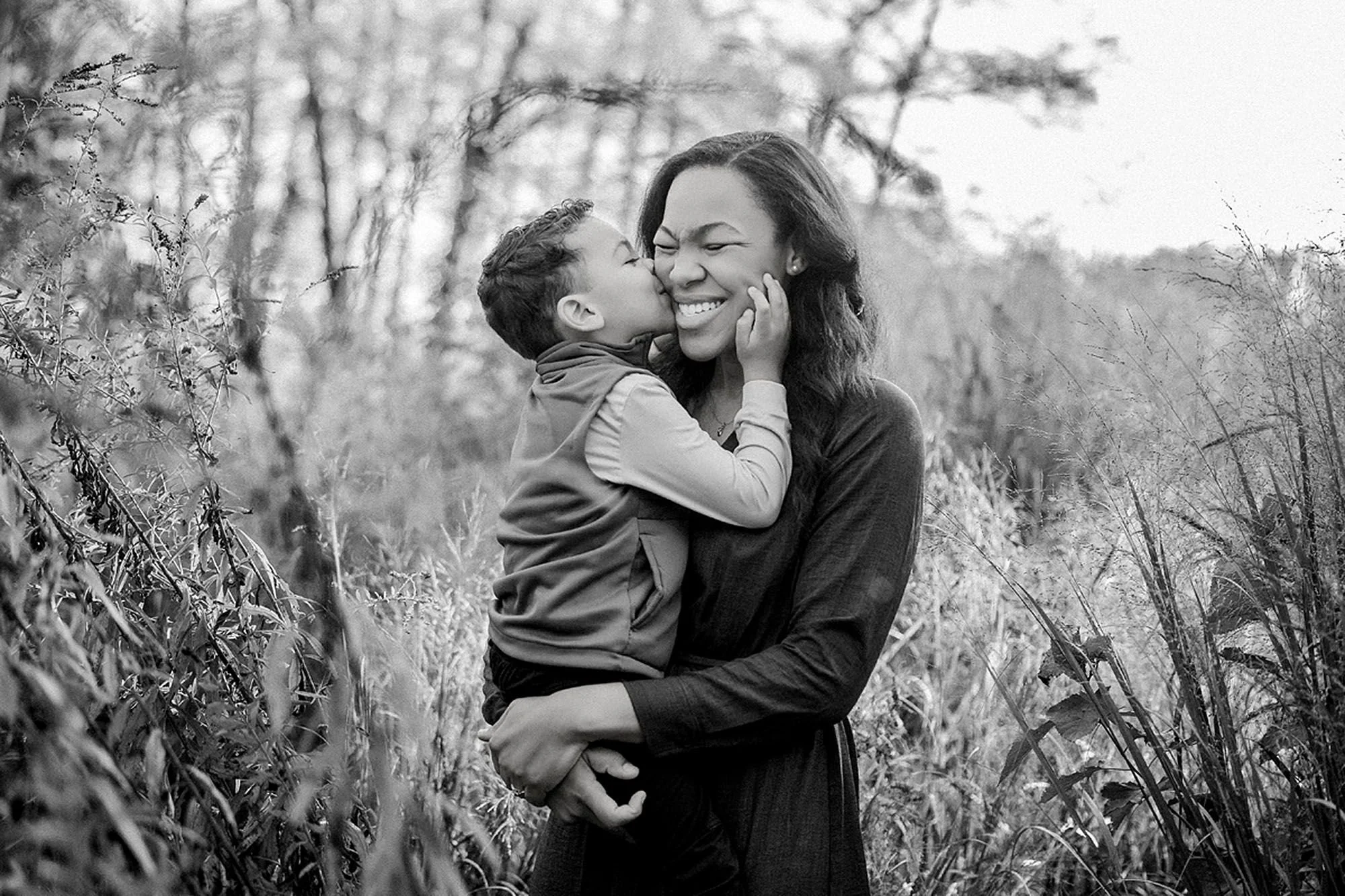 Black-and-white portrait of a young boy kissing his mother's cheek while she laughs, standing in tall grass while family photos capture a tender mother-son moment