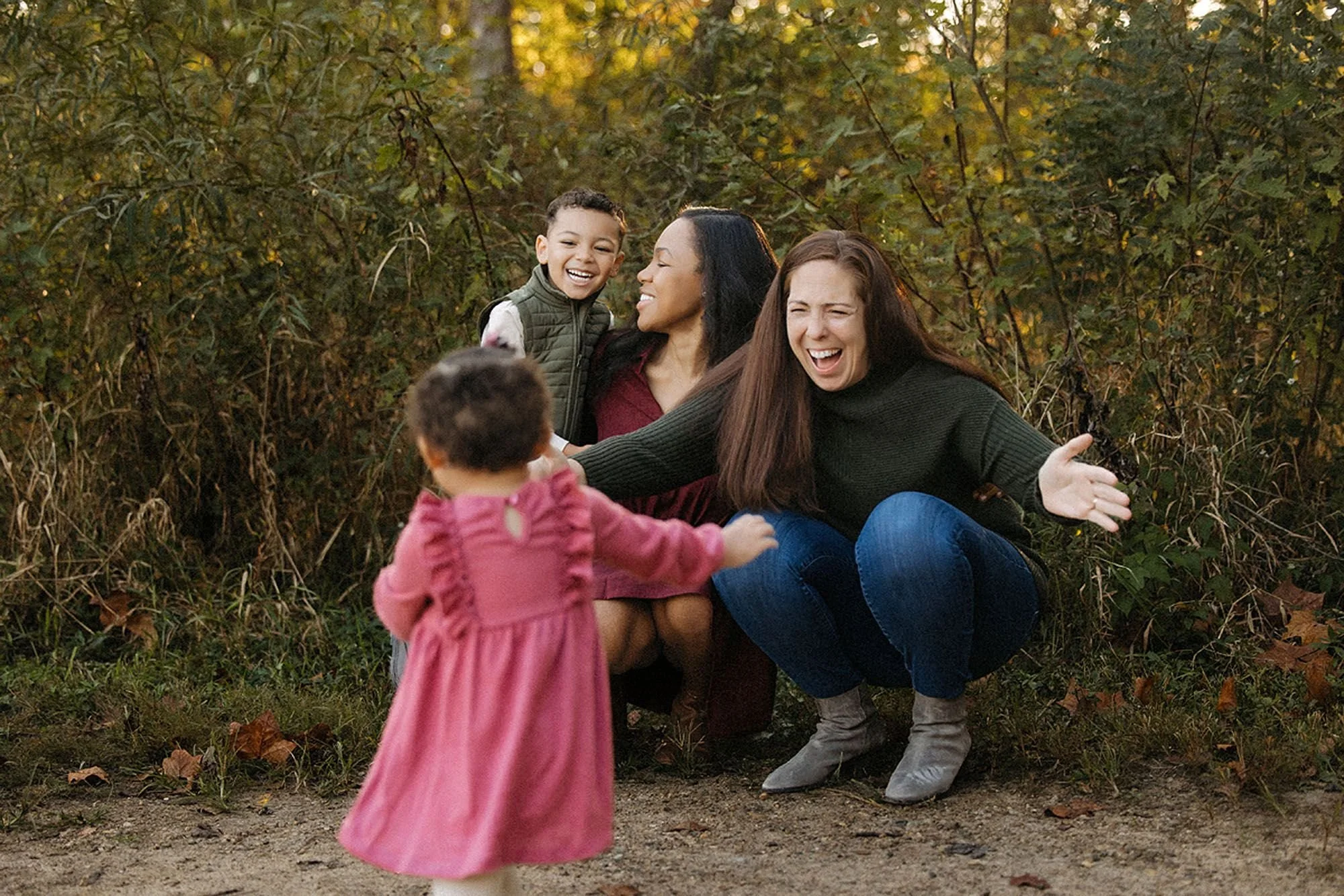 Two mothers crouching with open arms, laughing as their toddler runs toward them on a wooded path, capturing a joyful running-hug moment