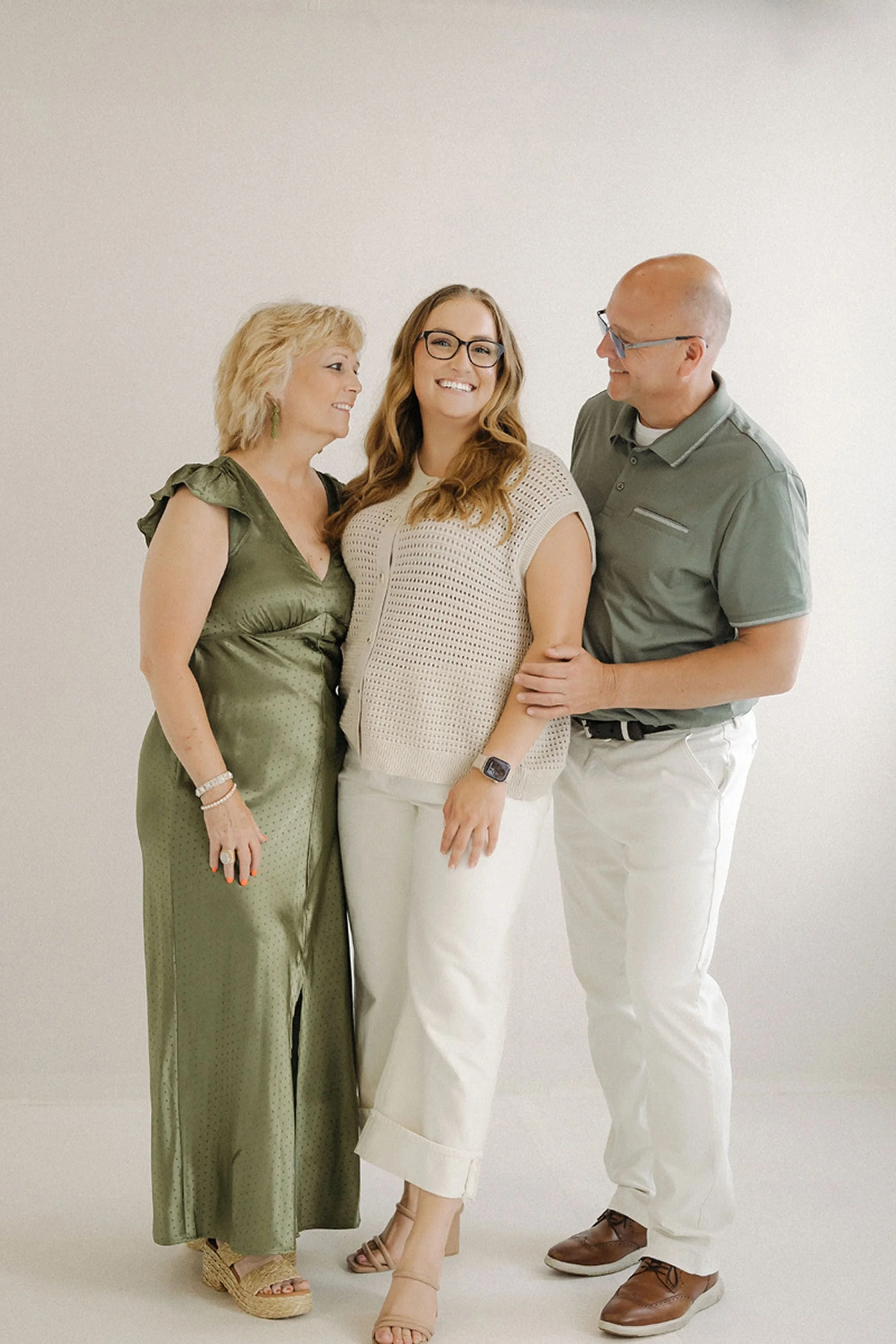 Smiling parents standing beside their adult daughter in a bright, minimalist studio, capturing a joyful family moment during Studio Family Photos at Studio Loft 18.